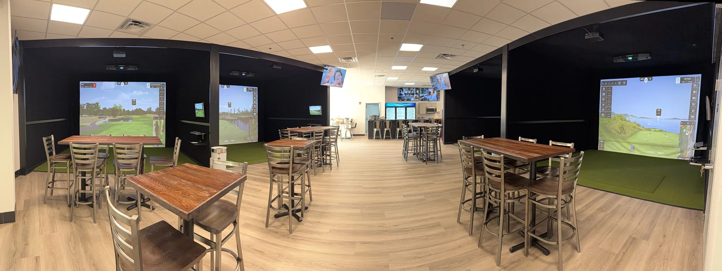 Indoor golf simulator lounge with multiple large screens displaying virtual golf courses, surrounded by tables and chairs for spectators or players, with a bar or refreshment area in the background.