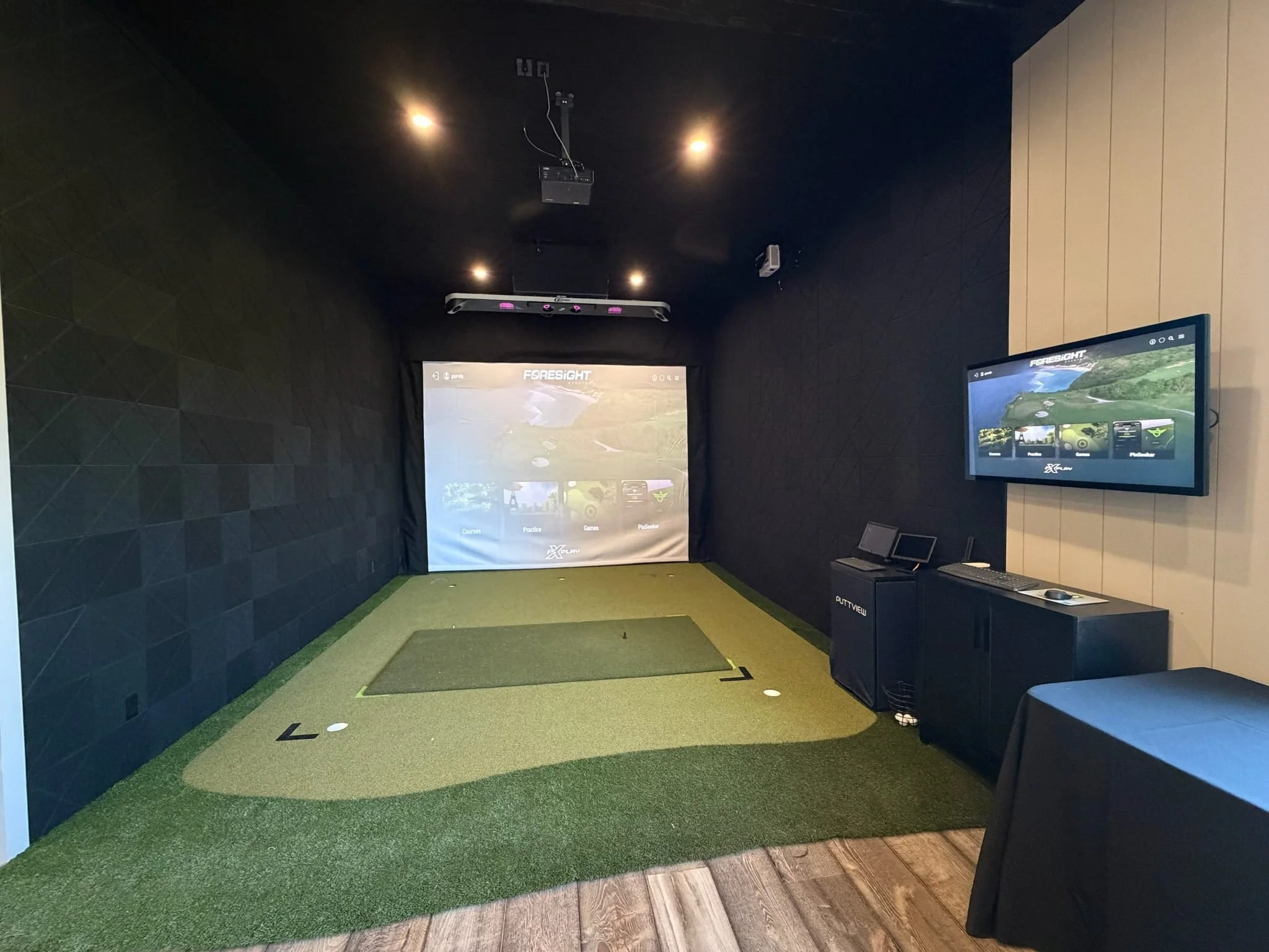 Indoor golf simulator room with a large screen displaying a virtual golf course, a smaller monitor, and golf mats on artificial turf floor.