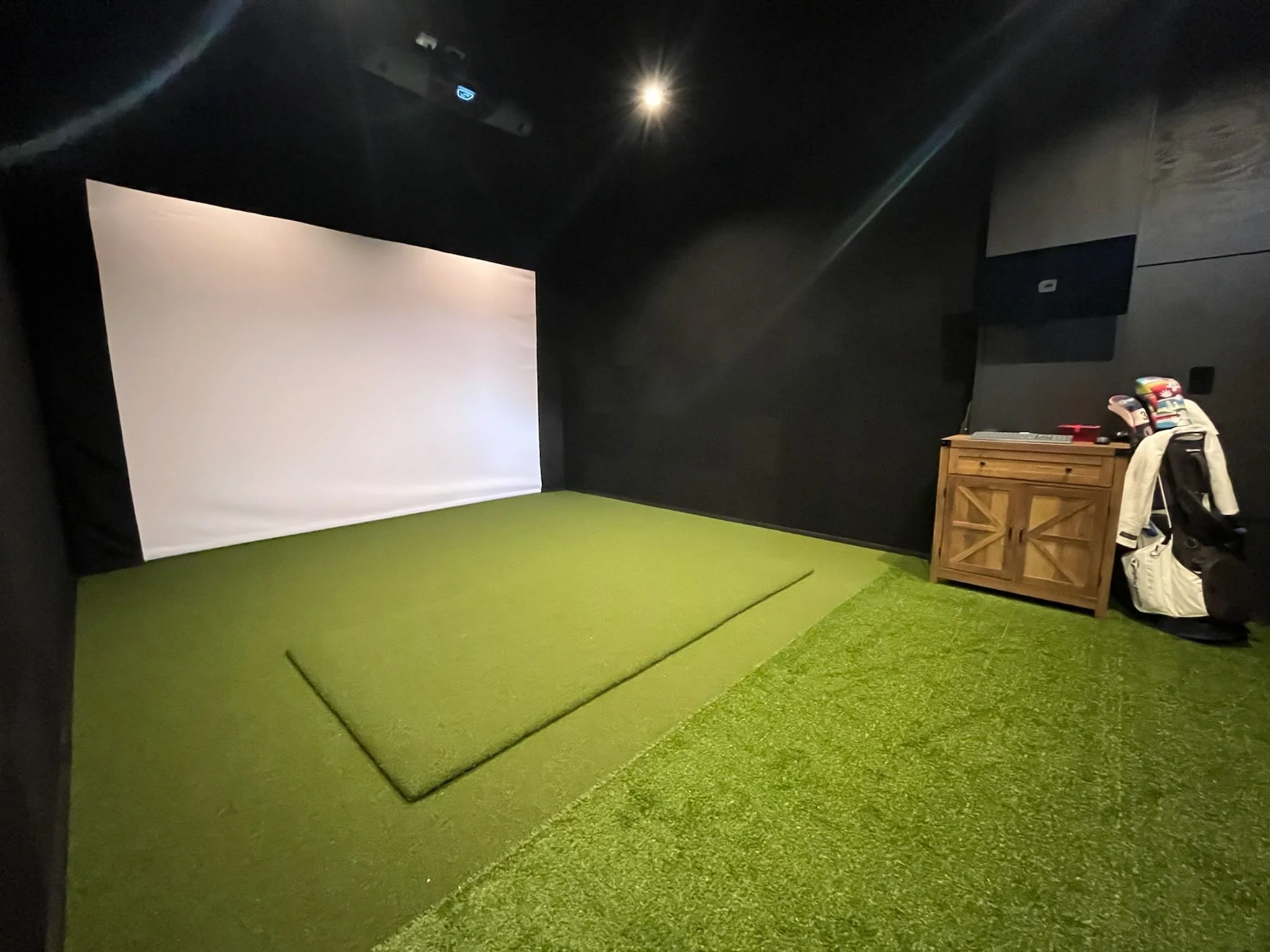 Indoor golf simulator room with a large white screen, green artificial turf floor, a wooden cabinet on the right, and a golf bag with clubs leaning against it.