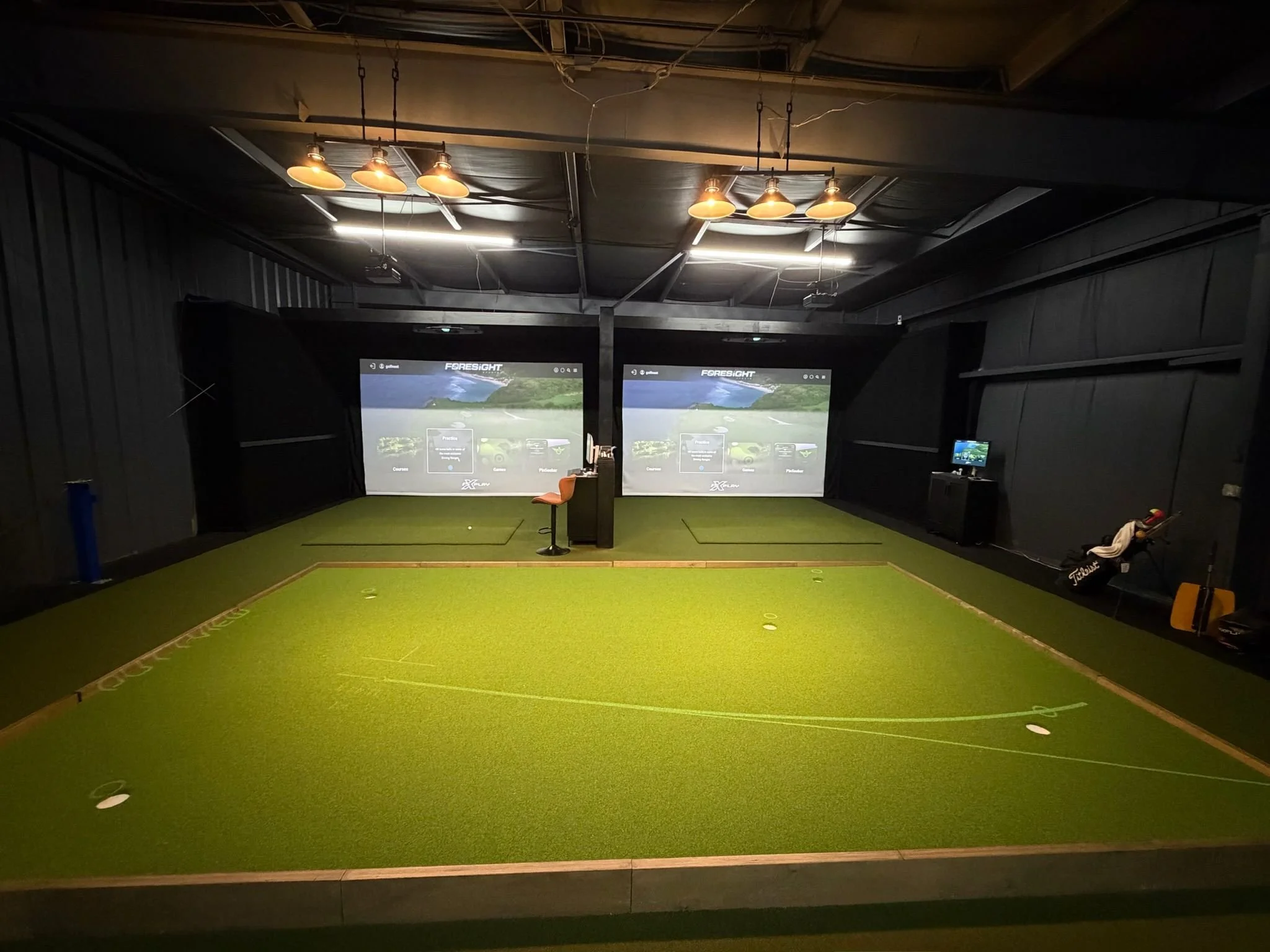 Indoor golf simulation room with artificial turf, two large screens displaying virtual golf courses, and golf equipment including a golf bag and a small TV on the right side.