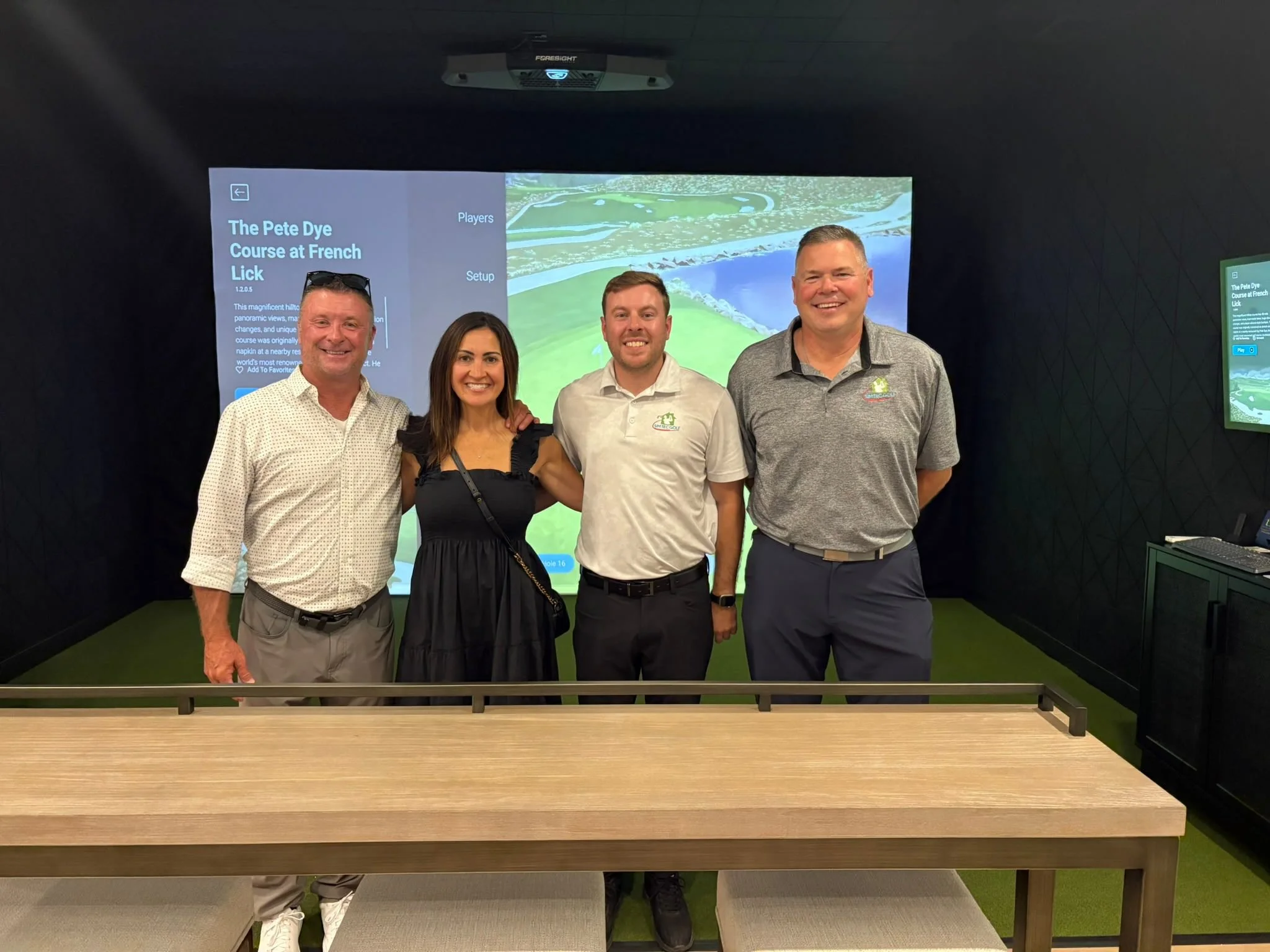Four people standing together indoors in front of a large display screen, smiling at the camera.