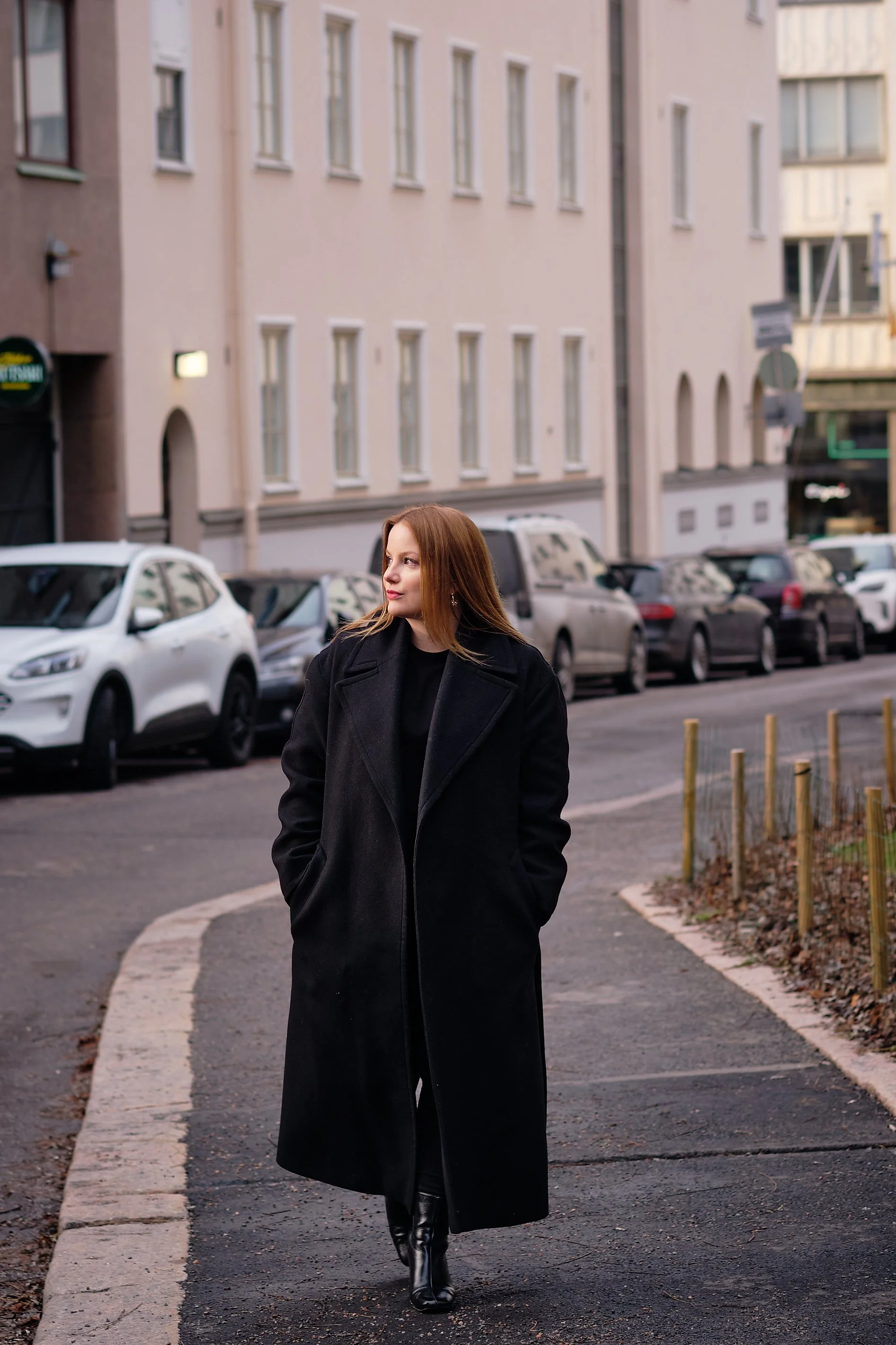 A woman with long red hair wearing a black coat and black boots walking on a city street.
