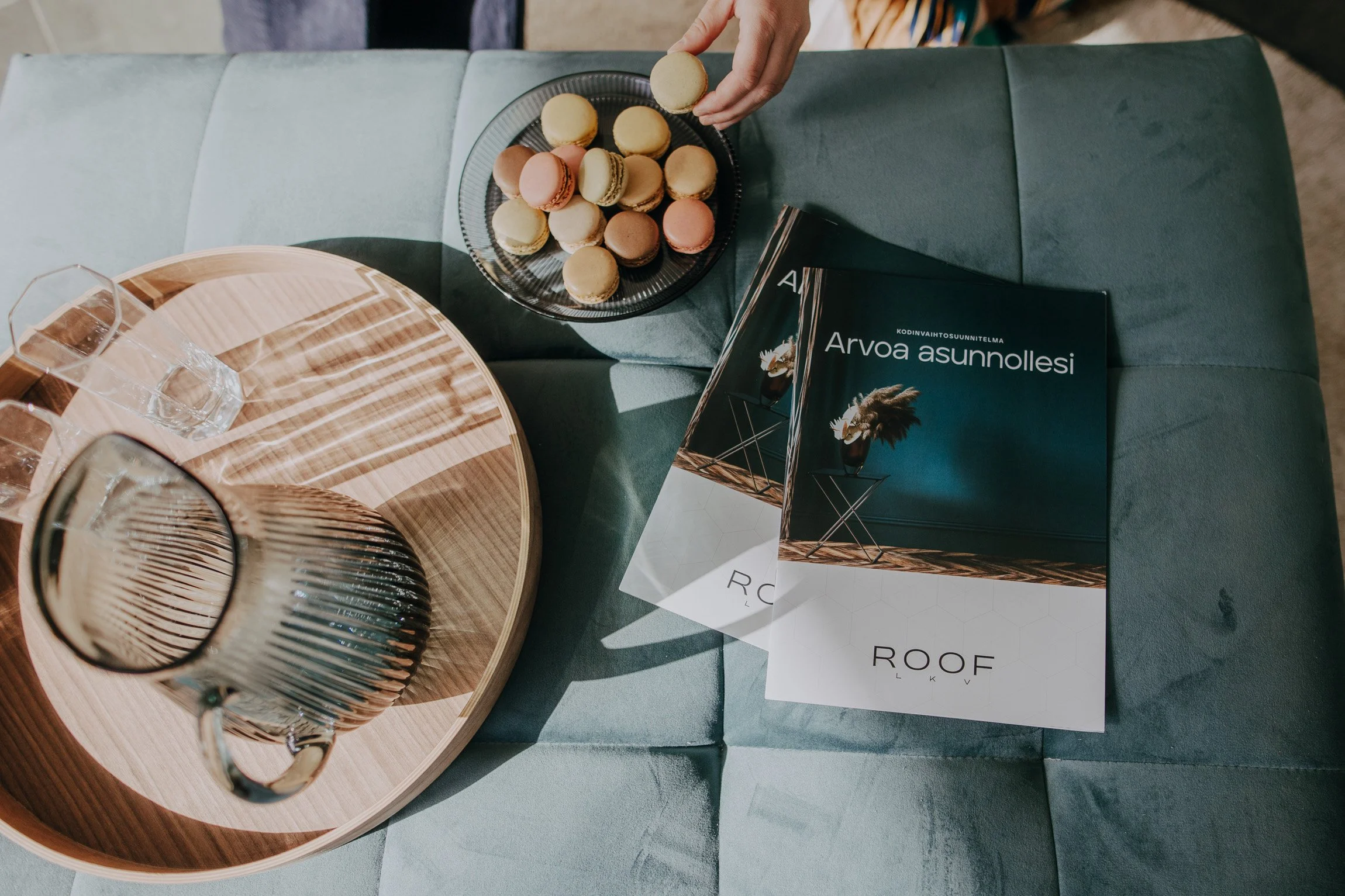 A person is reaching for a macaroon from a black plate filled with various pastel-colored macarons, placed on a green sofa beside a wooden tray with a glass of water and a magazine titled 'Arvoa asunnolsi'.