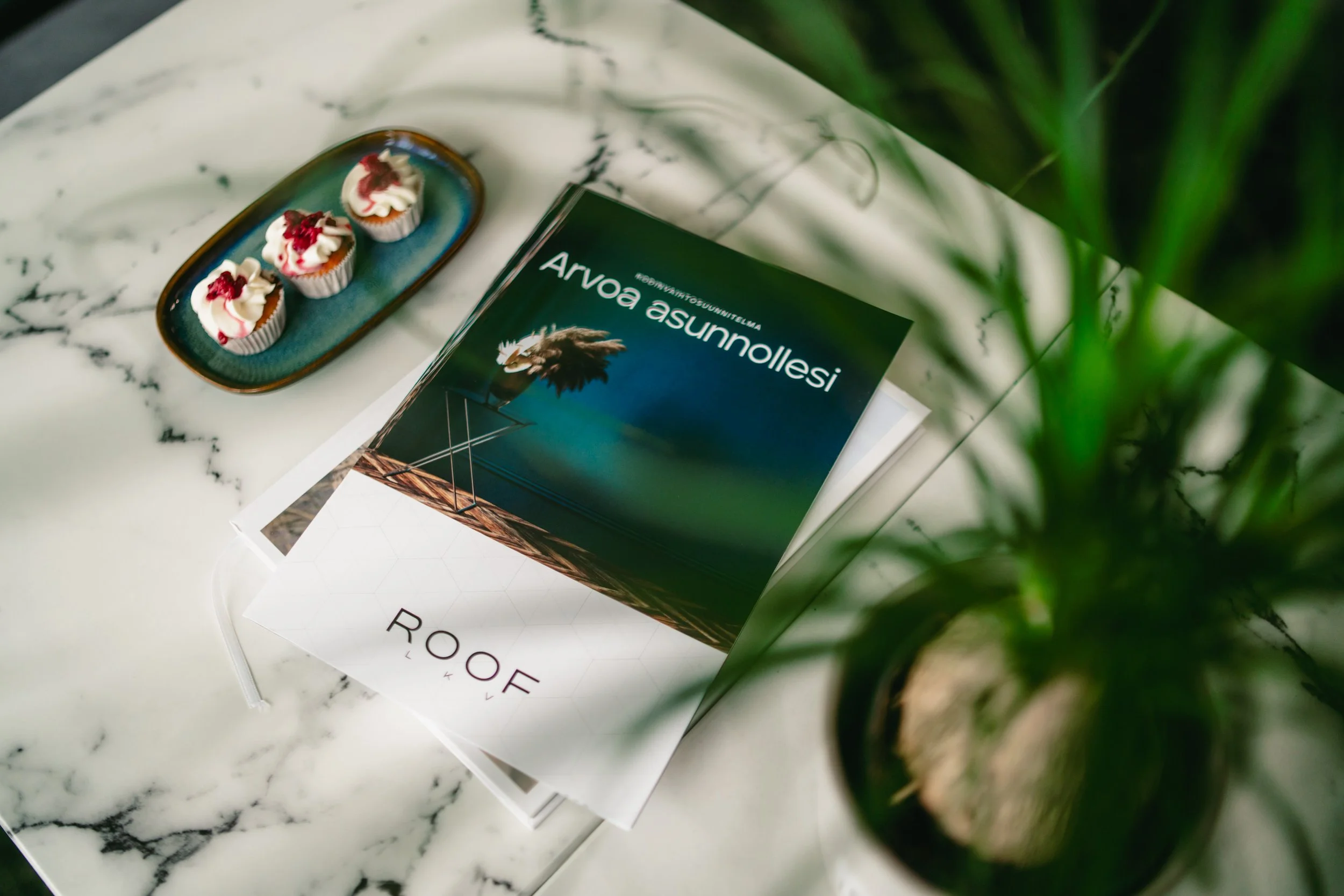 Three cupcakes with white frosting and red cherry toppings on a dark blue Ceramic plate, and a magazine about Arvoa asunnot on a white marble table with green plant in the foreground.