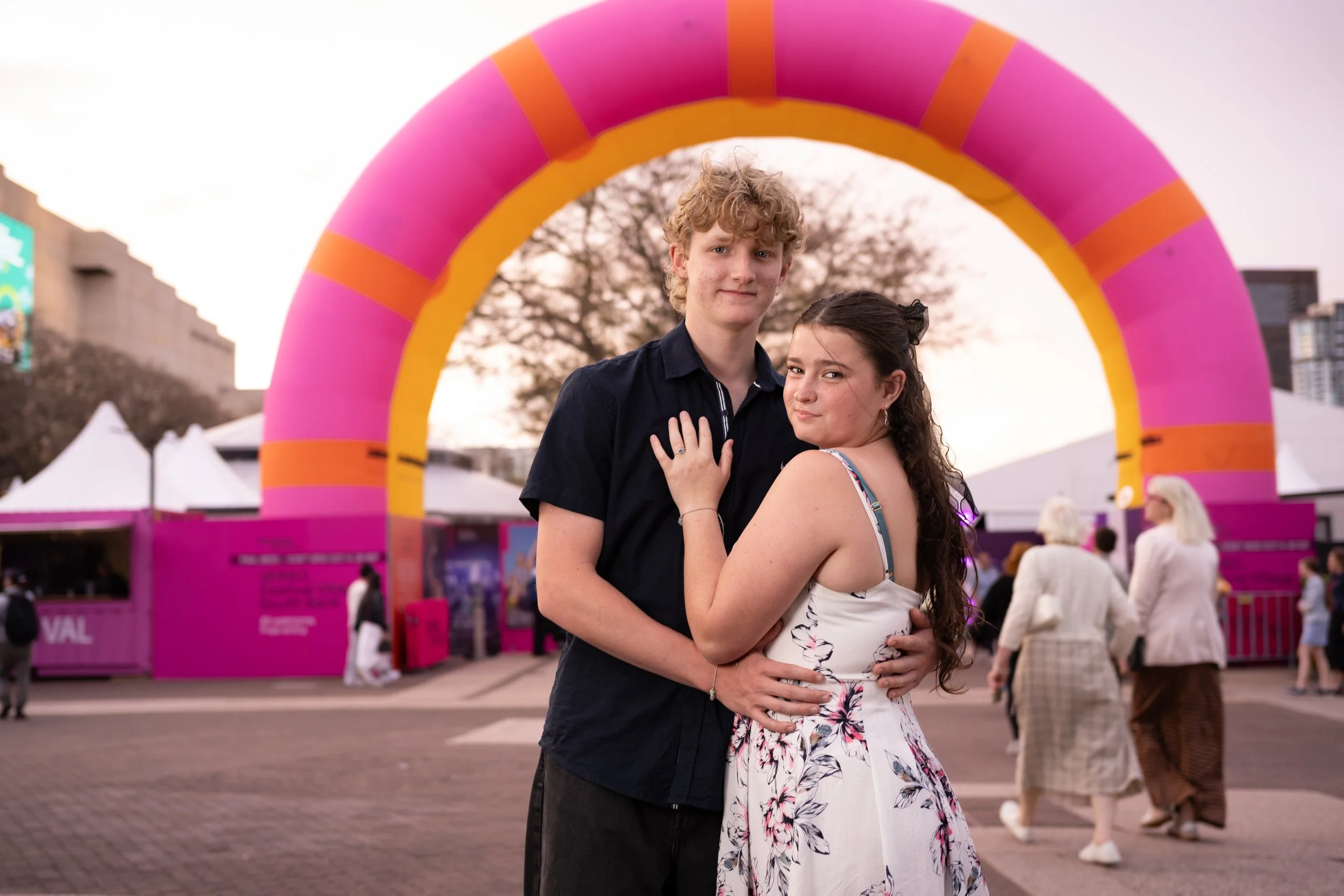 A young couple embracing each other at an outdoor event with a large pink and yellow inflatable arch in the background, and tents and people visible in the background.