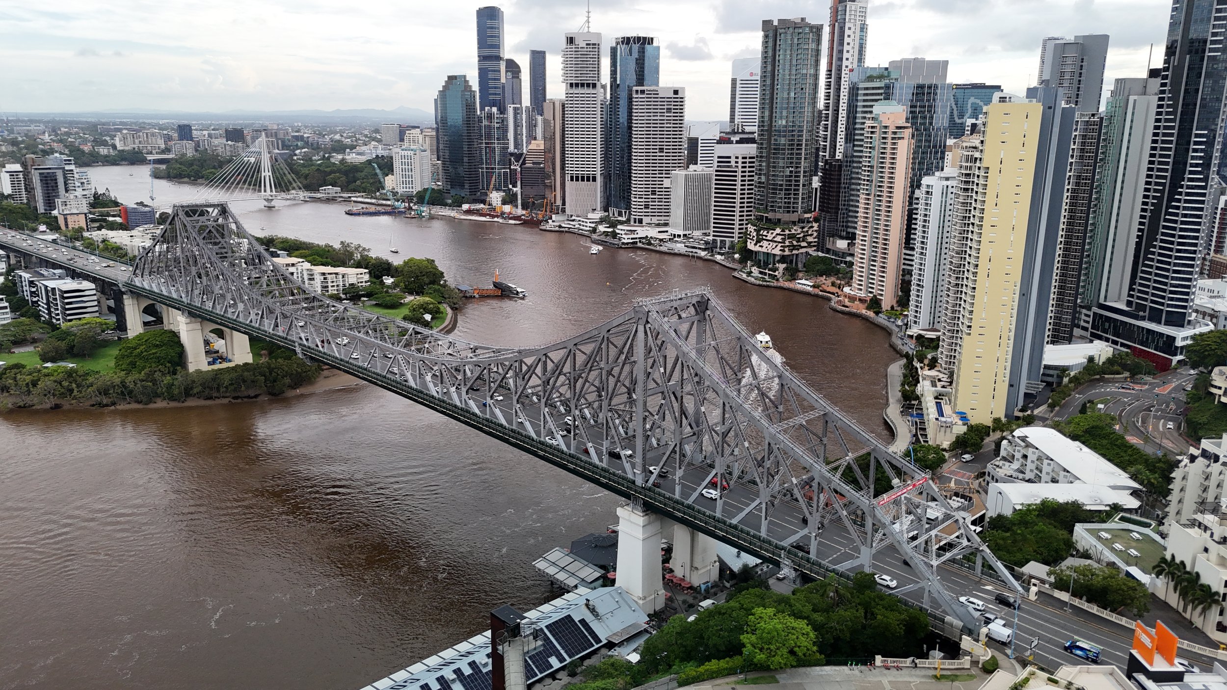An aerial view of Brisbane, Australia, showing the Brisbane River crossing through the city with a bridge, tall skyscrapers, and a mix of modern buildings.