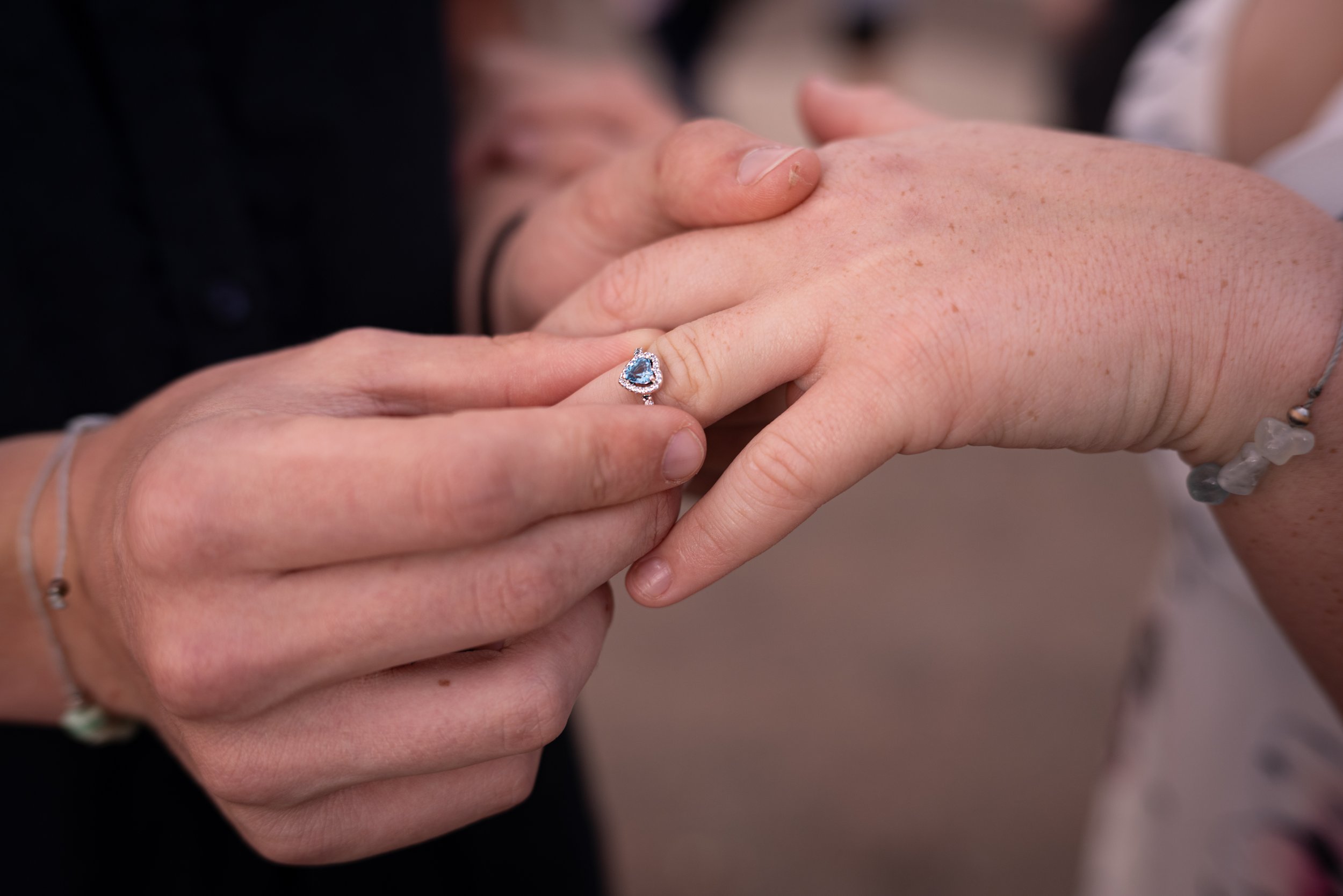 A close-up of two people holding hands, with one person wearing a heart-shaped ring with a blue gemstone. The person with the ring has fair skin and is wearing a beaded bracelet.