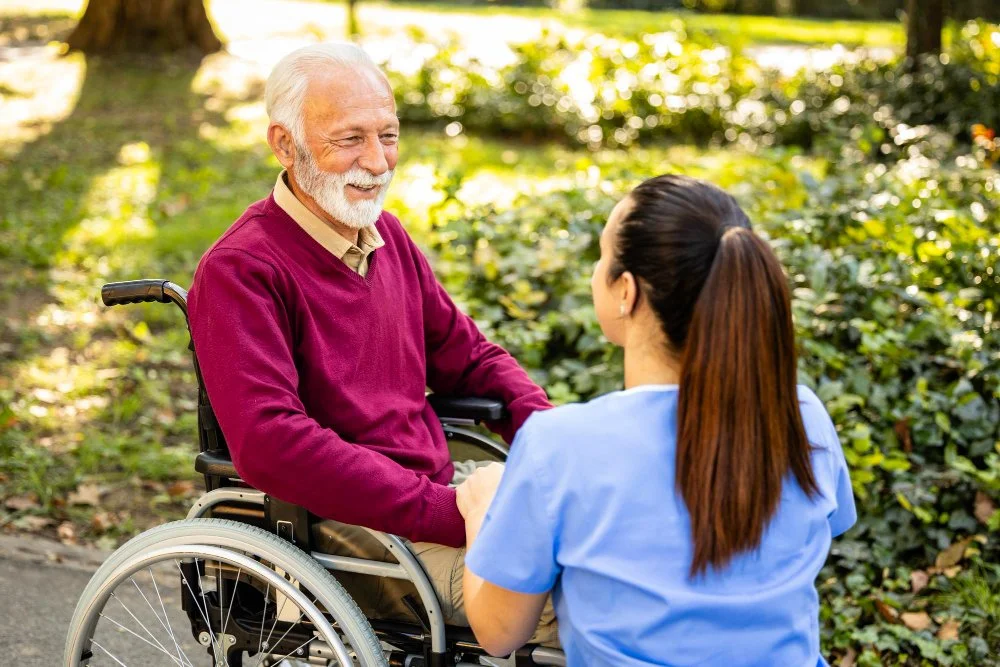 An elderly man with white hair and a beard smiling and holding hands with a young woman in a blue medical uniform outdoors in a park with green trees and bushes, sunlight filtering through the leaves.