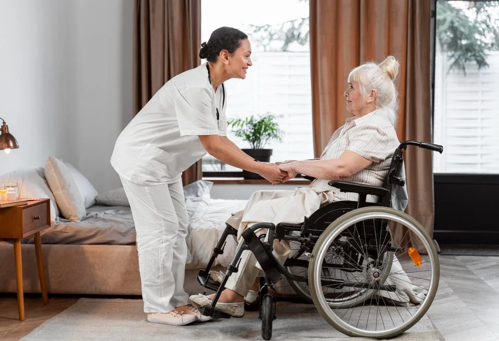 Nurse smiling and holding hands with an elderly woman in a wheelchair in a bedroom.