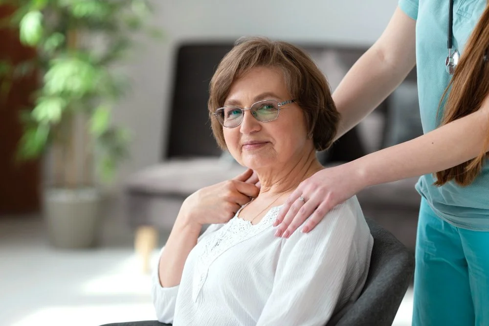 A senior woman sitting in a chair at a healthcare facility, receiving a shoulder massage from a nurse wearing scrubs.