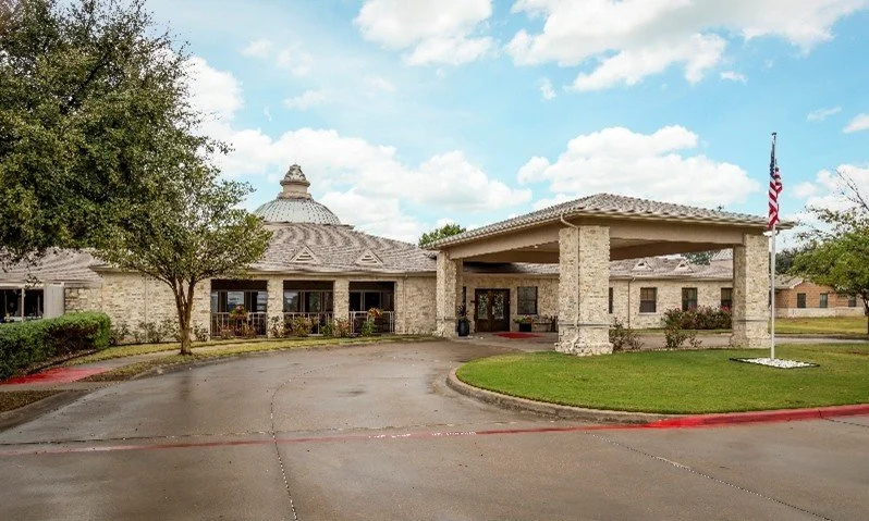 A stone building with a circular dome roof and a porte-cochère entrance, surrounded by well-maintained lawns and trees, with an American flag on a pole nearby.