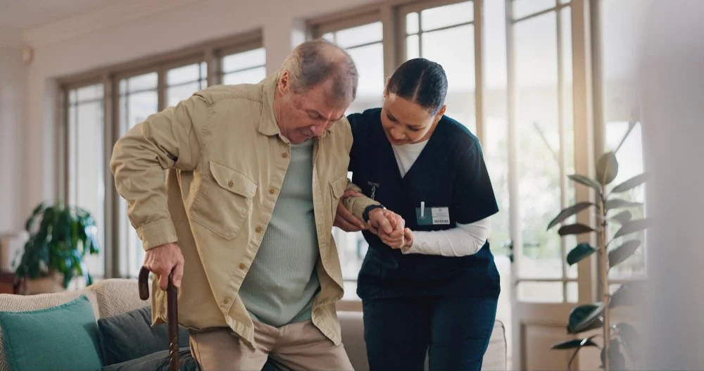 A caregiver assisting an elderly man with walking in a bright, modern living room.