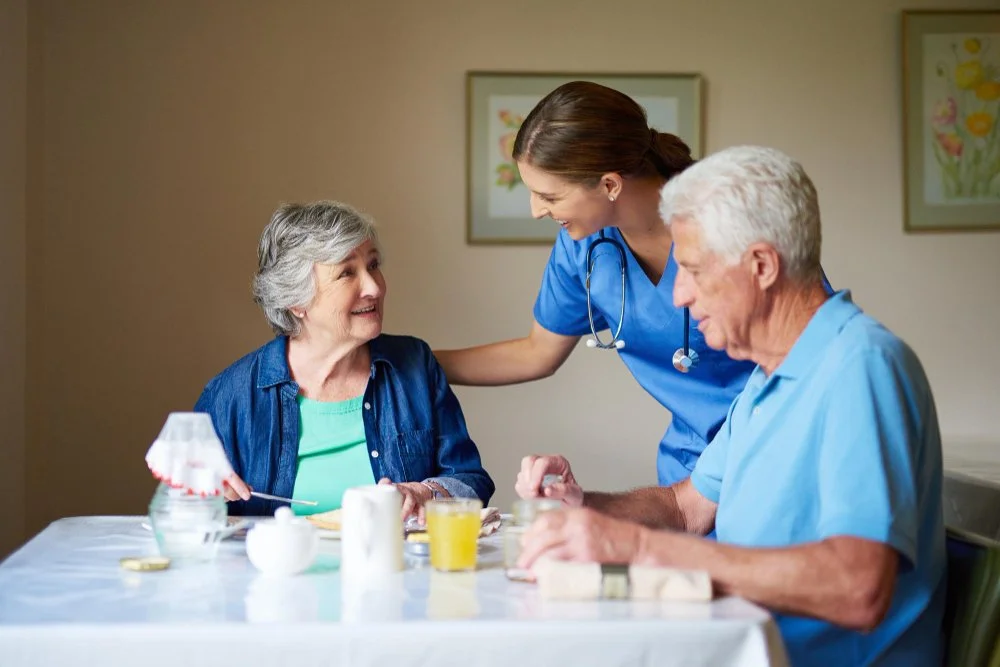 A nurse in blue scrubs smiling and talking to an elderly woman and man, who are sitting at a table with food and drinks, in a room with framed flower pictures.