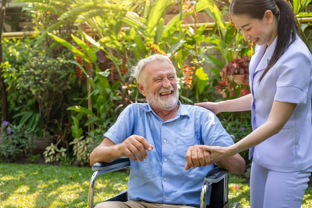 An elderly man with a beard smiling and in a wheelchair, interacting happily with a young female medical caregiver in a white uniform, outdoors in a garden.