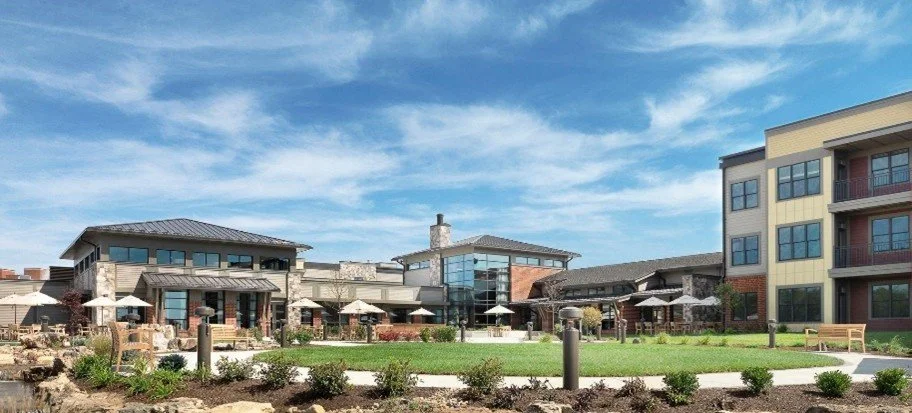 Outdoor view of a modern apartment complex with a landscaped courtyard, several patio areas with umbrellas, and a blue sky.