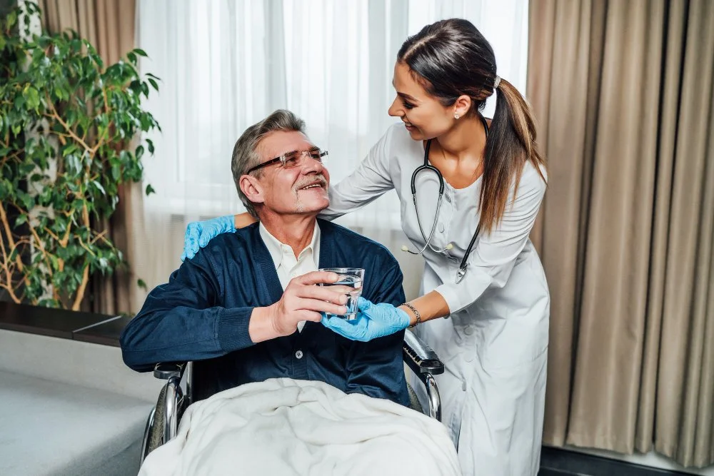 A female healthcare professional wearing a white coat, stethoscope, and gloves giving a glass of water to an elderly man in a wheelchair, smiling at each other in a medical setting.