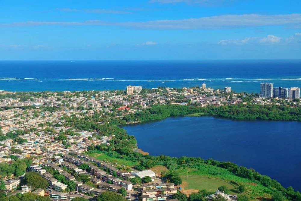 Aerial view of a coastal city near a large lake with an ocean in the background, showing urban areas, greenery, and bodies of water under a partly cloudy sky.
