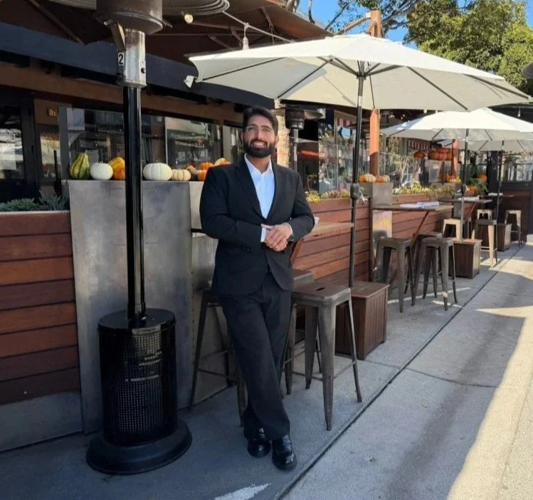 A man in a dark suit standing outdoors at a cafe or restaurant with white umbrellas, wooden tables, and chairs, and decorative pumpkins on a ledge behind him.