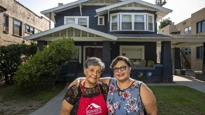 Two women standing in front of a house, smiling. One wears a black lace top and red apron, and the other wears a floral sleeveless top and glasses.