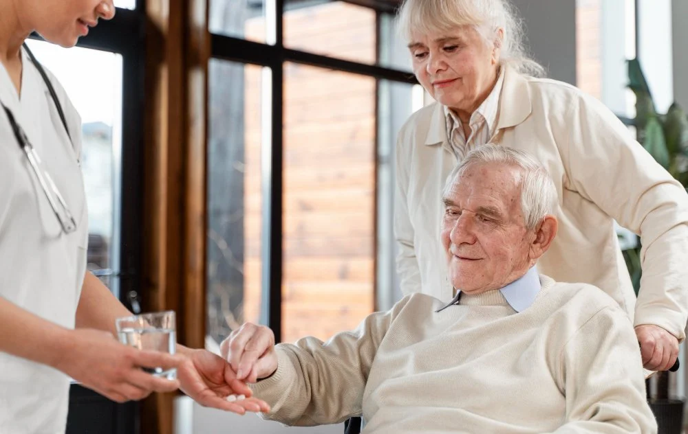 A healthcare professional handing a glass of water to an elderly man, while an older woman looks on in a healthcare setting with large windows.