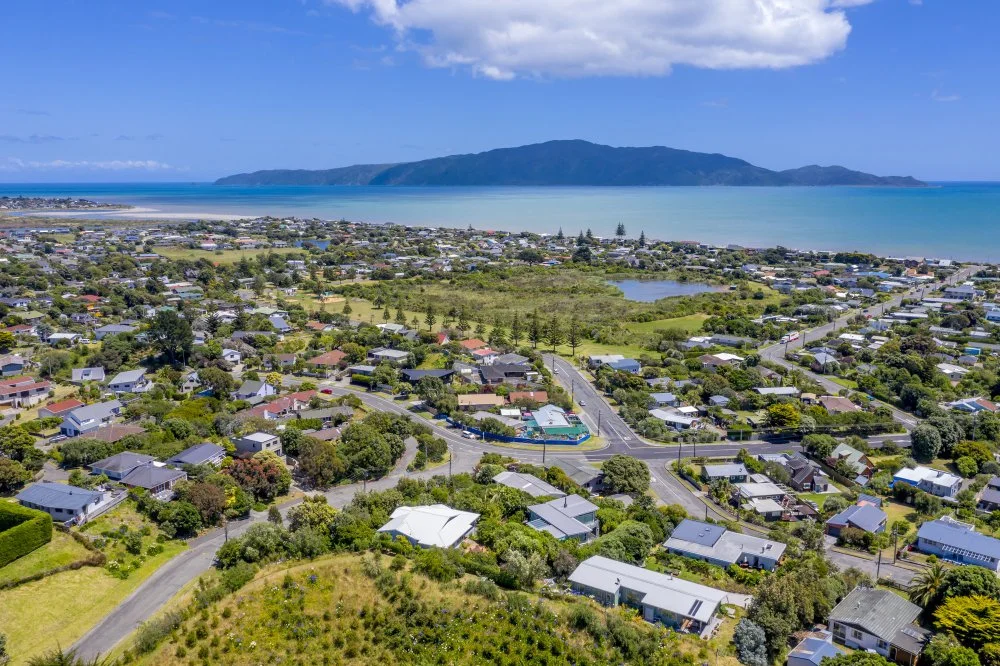 Aerial view of a coastal town with houses, green spaces, and a bay, with a mountain in the background under a partly cloudy sky.