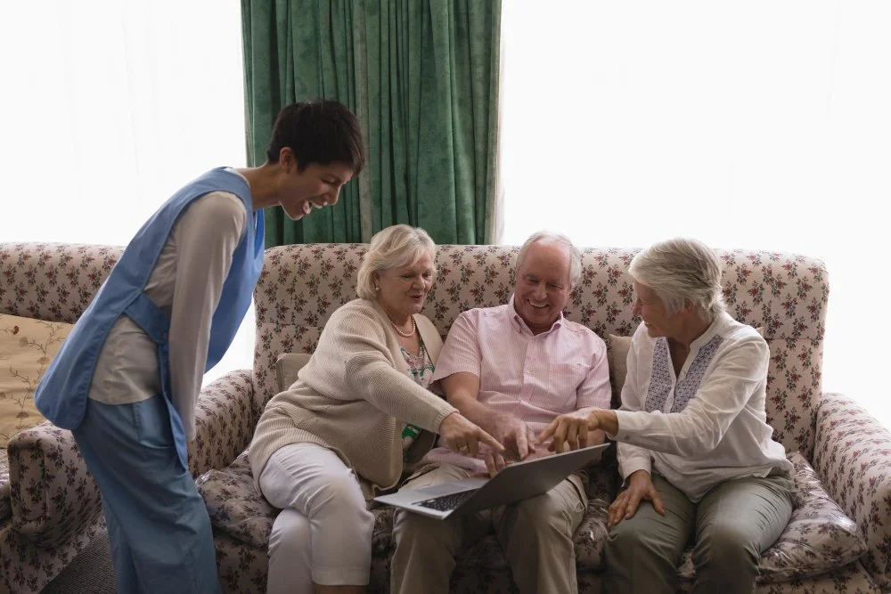 Four older adults and a younger caregiver looking at a laptop together on a floral couch in a bright room with green curtains.