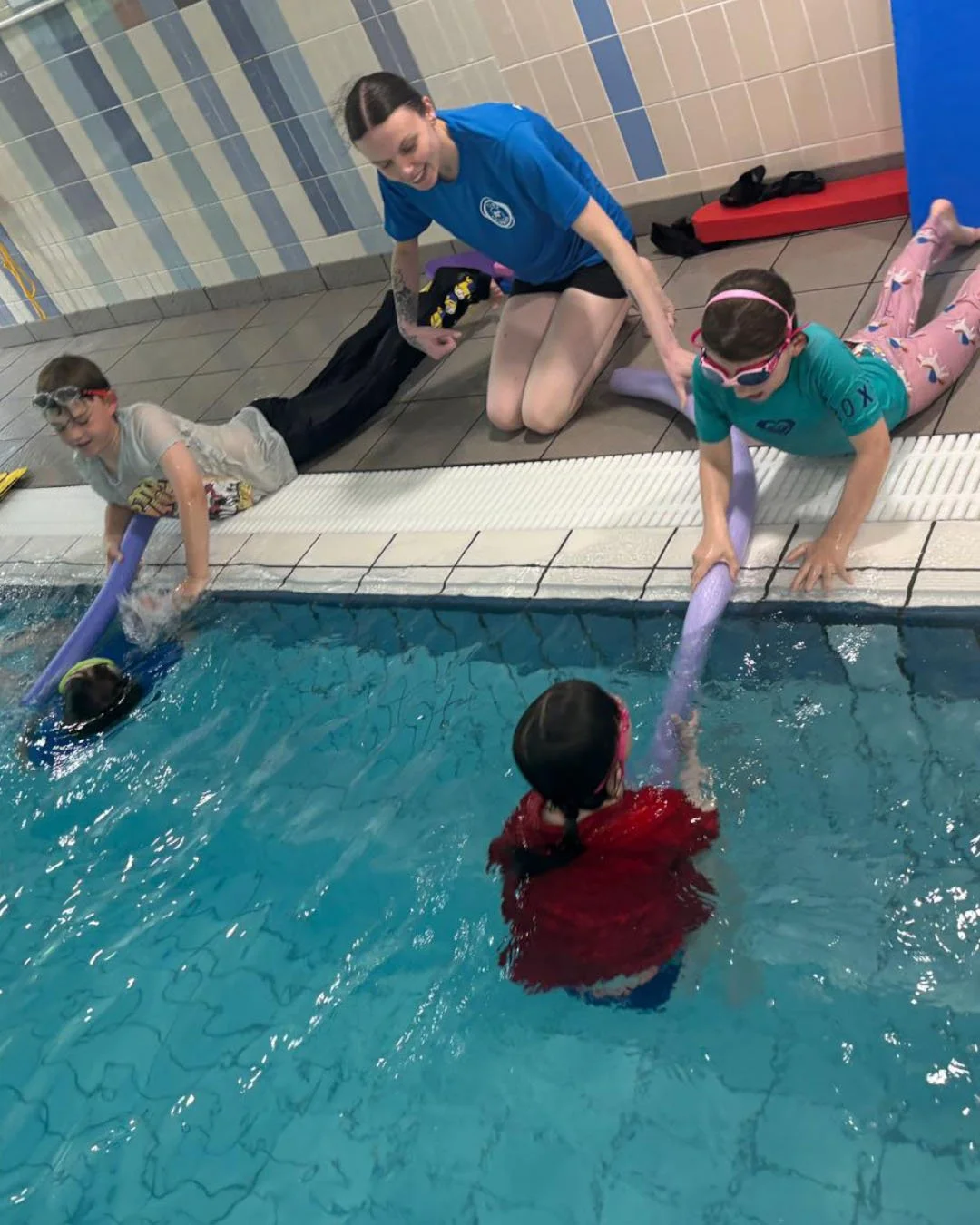 Swim instructor helping two young girls learn to swim in an indoor pool, with a child practicing with a pool noodle in the water.