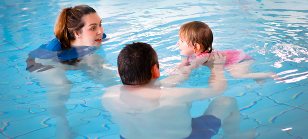 A woman teaches a young girl to swim in a swimming pool with a man swimming alongside them.