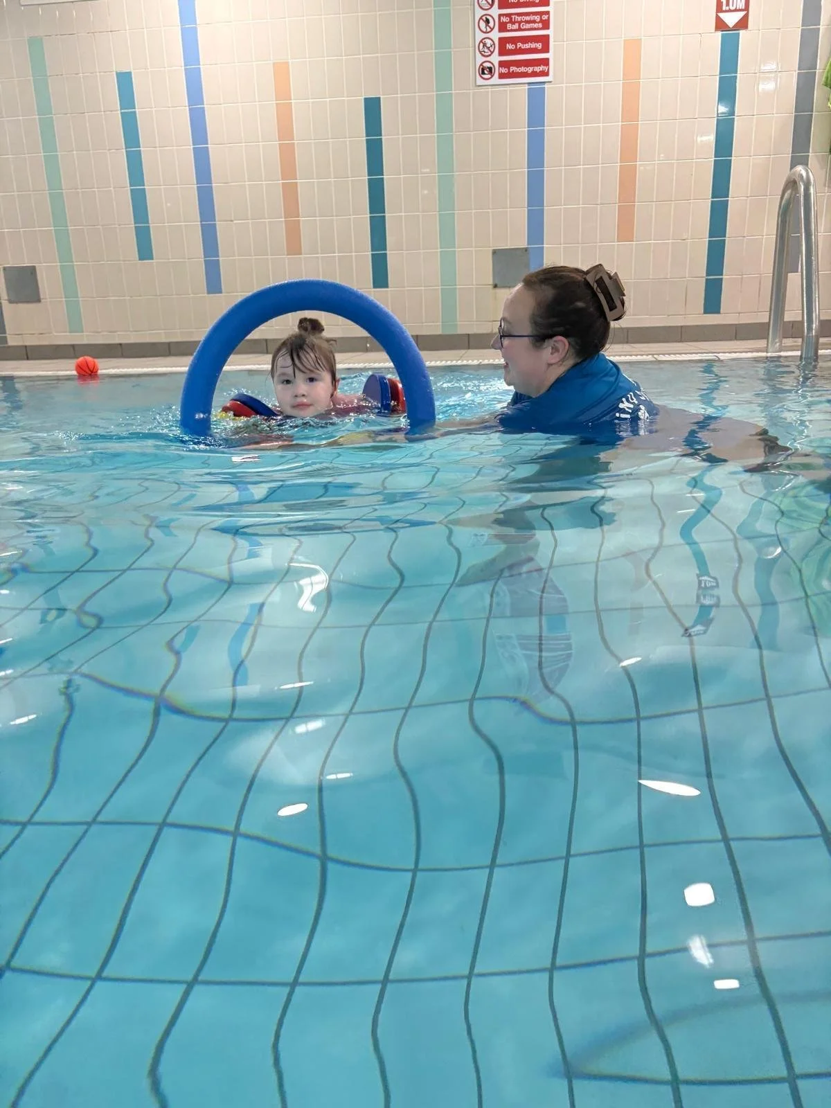 A young child with dark hair tied in a bun in a swimming pool with a blue pool float, accompanied by an adult woman with glasses and dark hair in a bun, near the pool's ladder. There are colorful tiles on the wall behind them and a sign with pool rules.