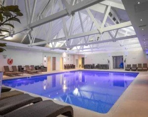 Indoor swimming pool with lounge chairs along the sides and a high, white ceiling with exposed beams.