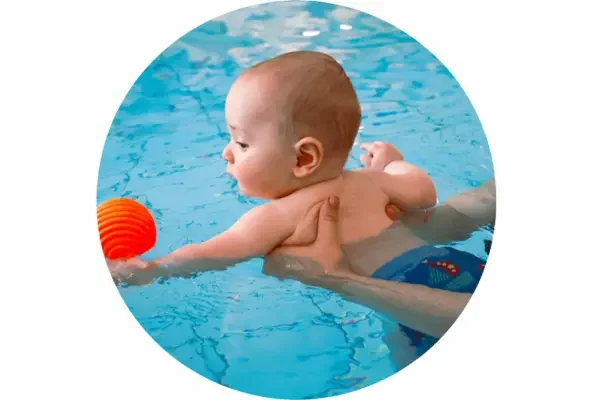A baby swimming in a pool, reaching for an orange ball.