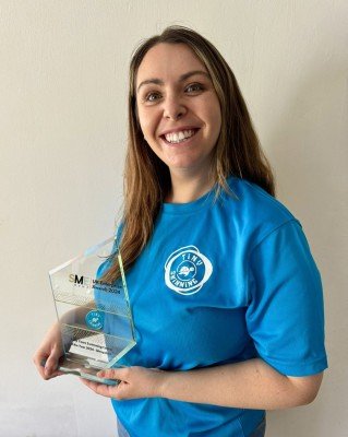 A woman with long brown hair in a blue shirt holding a clear award trophy, smiling against a plain wall.