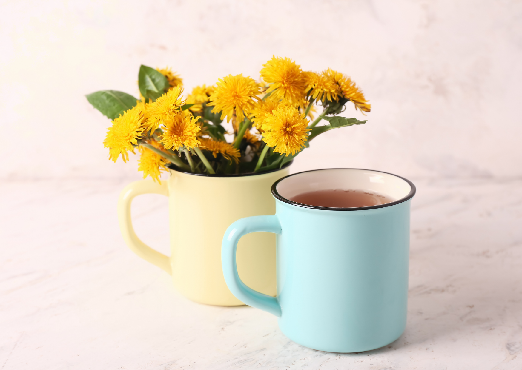 Zwei bunte Tassen, eine mit gelben Blumen und grünem Blatt, die andere mit einem warmen Getränk, stehen auf einer weißen Oberfläche.
