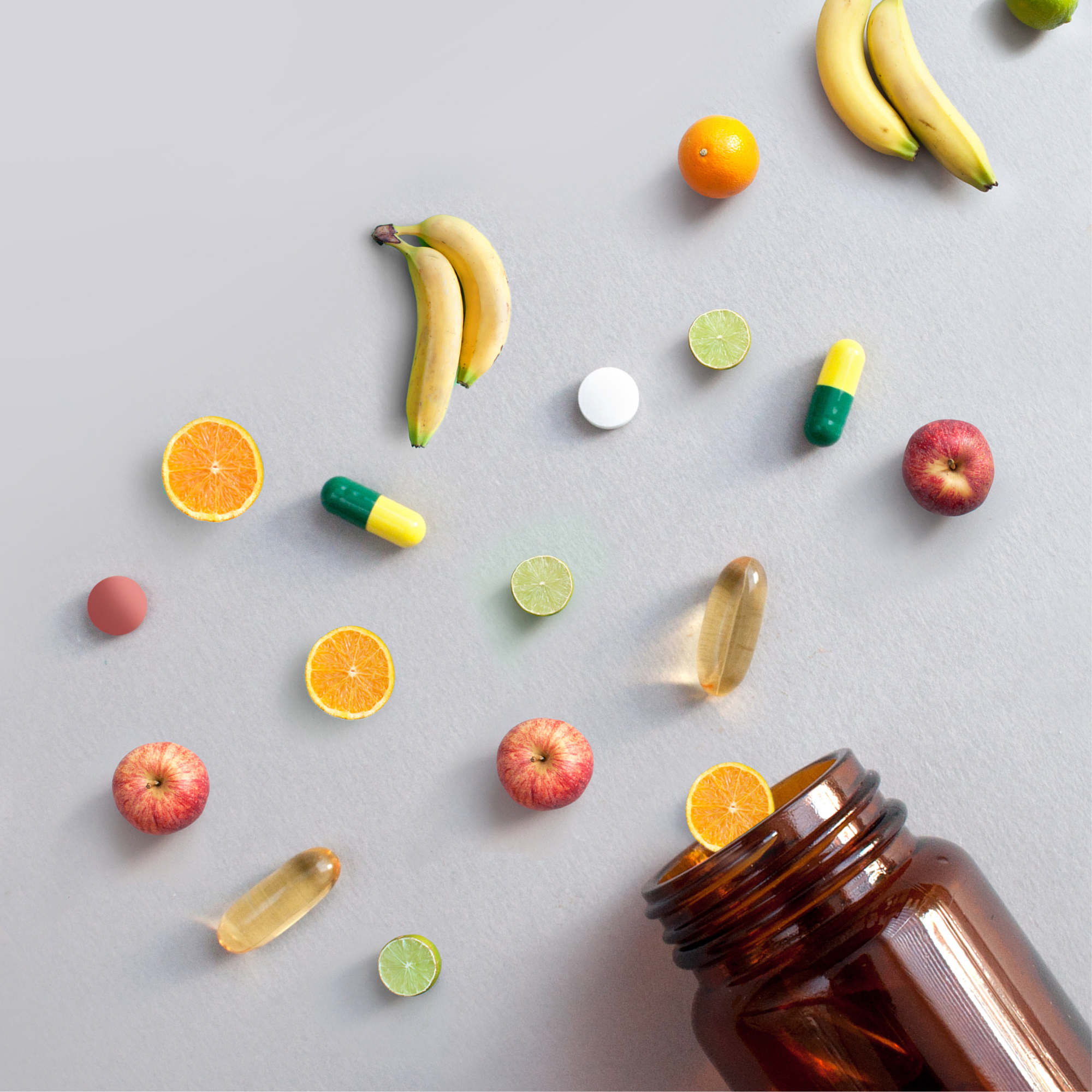 Verschiedene Früchte wie Bananen, Orangen, Limetten und Äpfel sowie einige Pillen und eine Medizinflasche auf einem weißen Hintergrund.