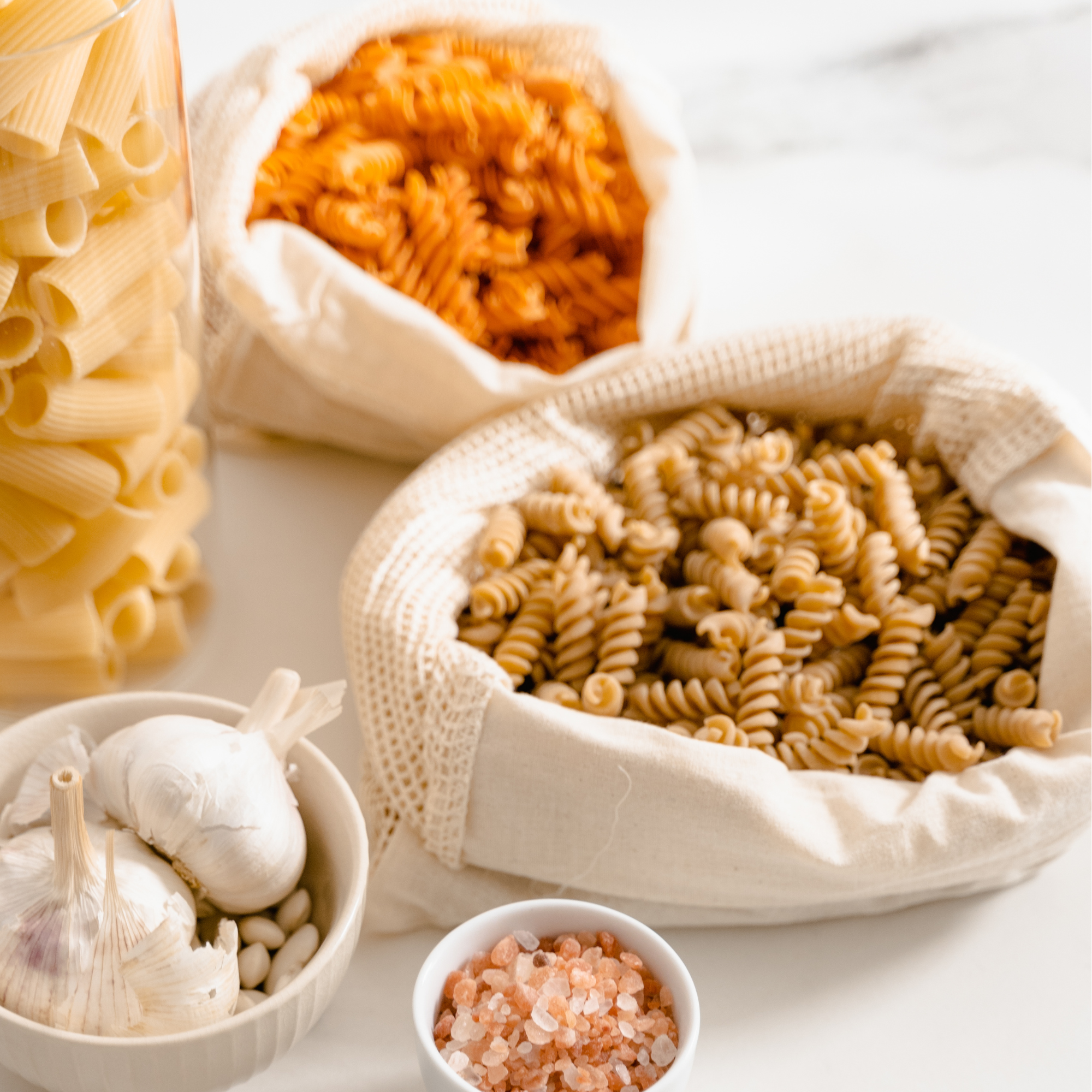 Verschiedene Sorten ungekochter Pasta, Knoblauch, Salz und Salzflocken in kleinen Schalen auf weißem Hintergrund.