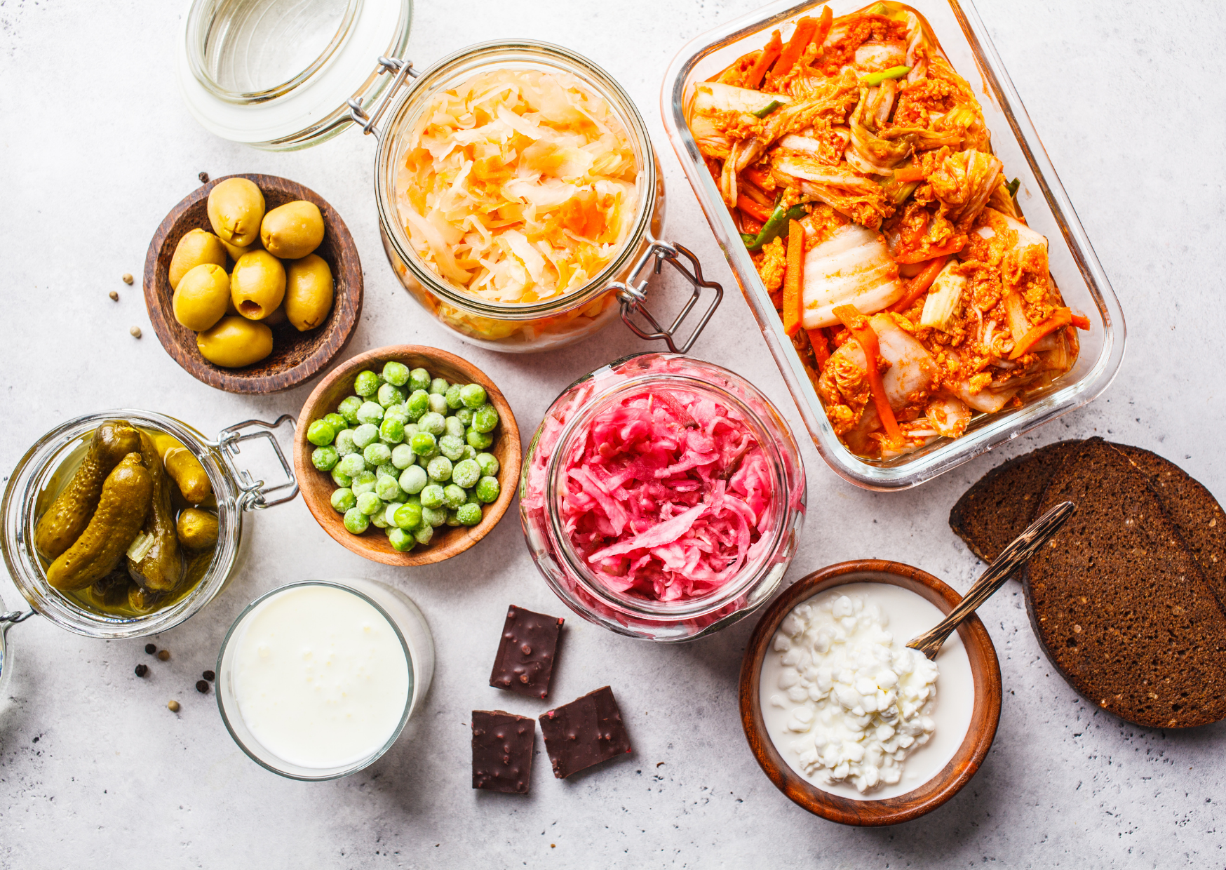 Verschiedene eingelegte Lebensmittel, Sauerkraut, Gurken, Erbsen, rotes Kraut, und andere Zutaten in Gläsern, neben einem dunklen Kuchen auf einer hellen Oberfläche.
