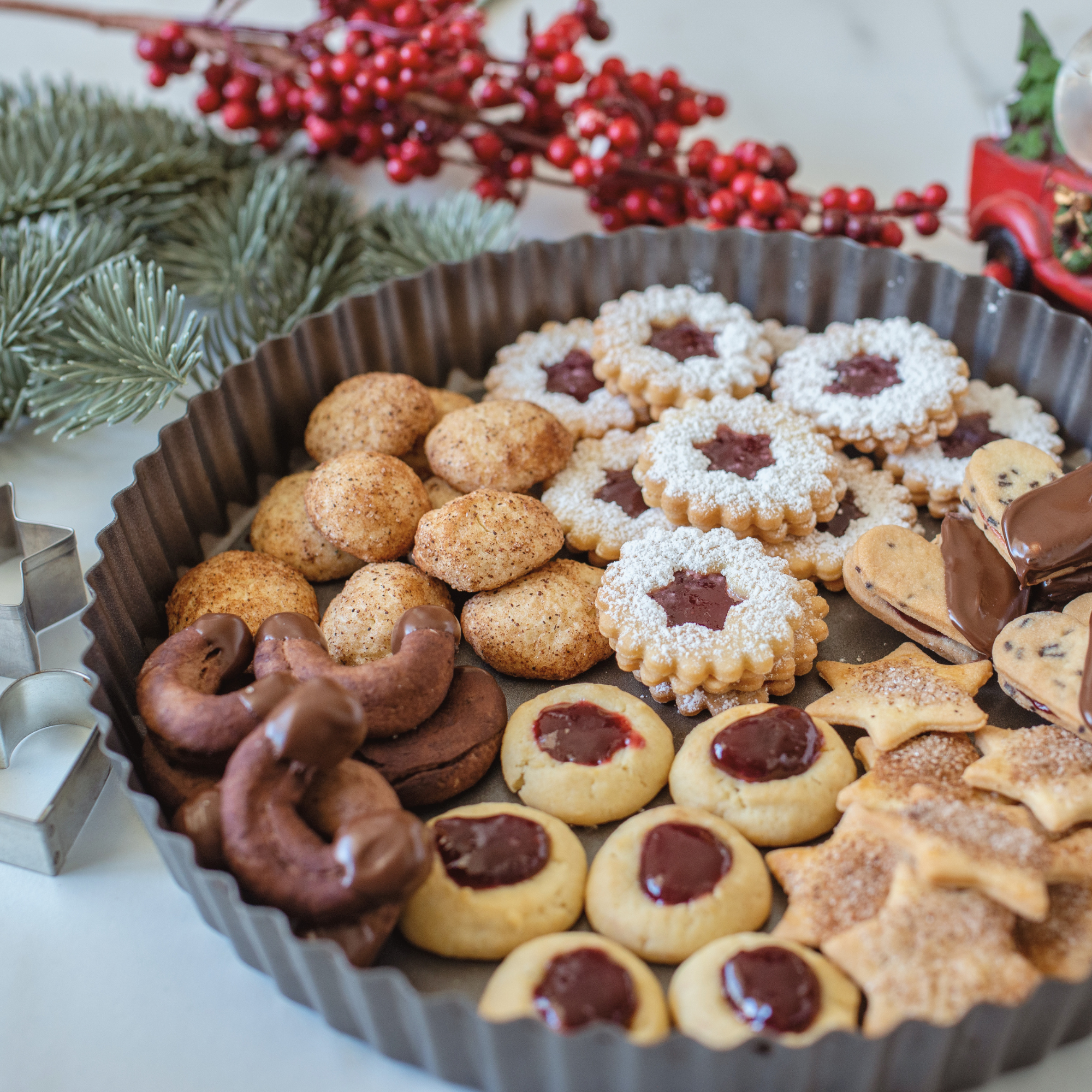 Verschiedene Kekse auf einem rechteckigen Teller, darunter Linzer Augen, Kokosmakronen und Schokoladenkekse, mit Tannenzweigen und roten Beeren im Hintergrund.