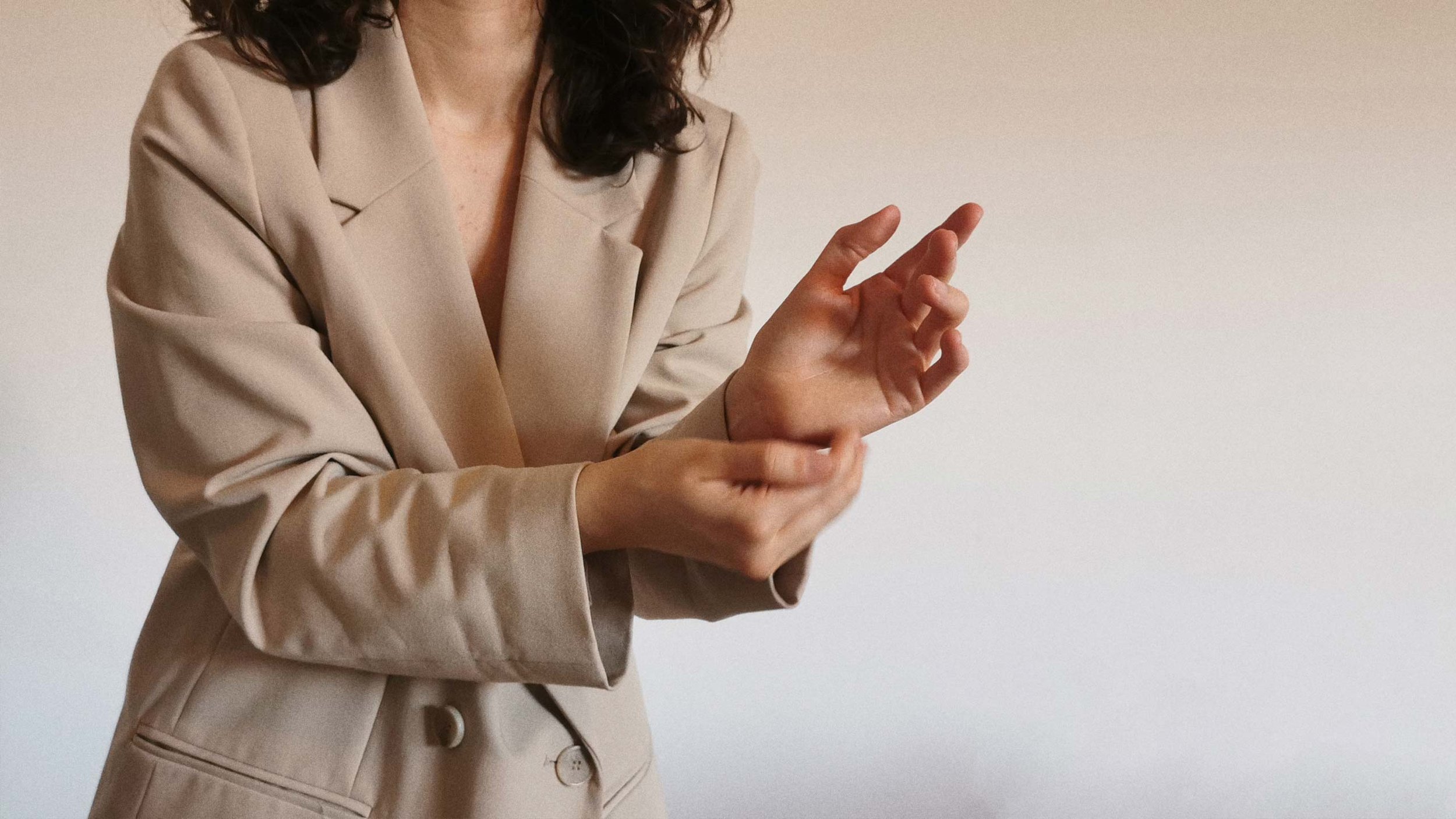 A woman in a beige blazer is gesturing with her hands in front of a plain light background.
