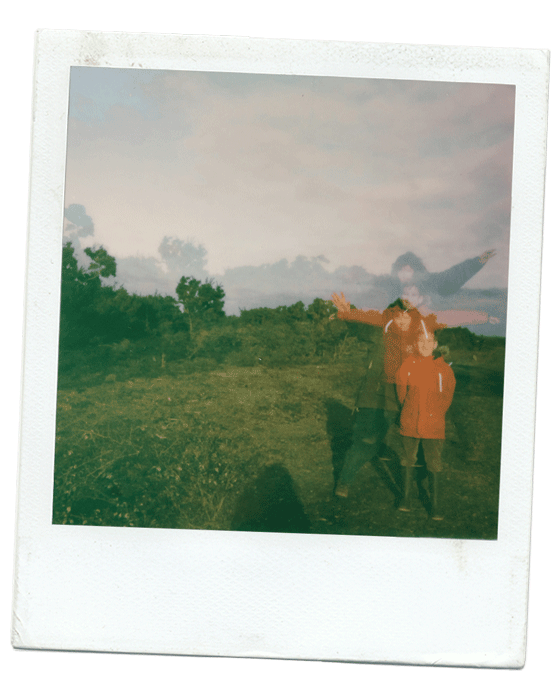 A blurred Polaroid photo of two children standing outdoors in a grassy field, with trees and a cloudy sky in the background.