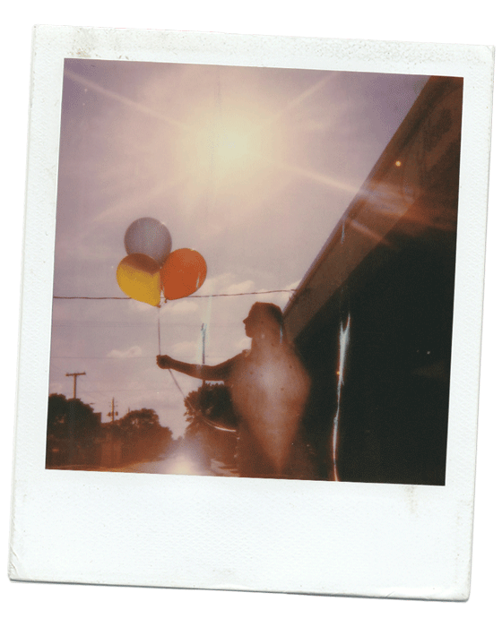 A person holding three balloons—white, yellow, and orange—standing outdoors with a bright sun overhead and a partly cloudy sky.