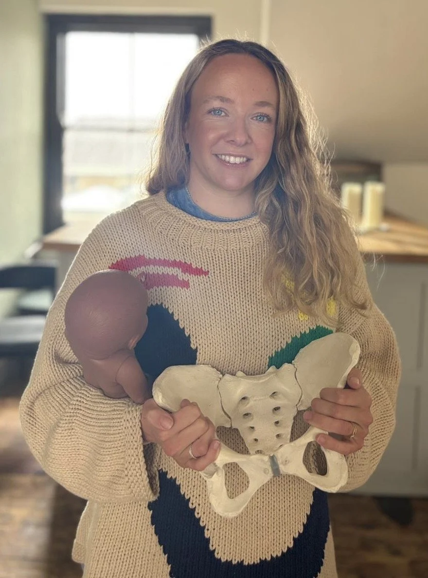 Woman smiling, holding a large pelvic bone model and a small model of a fetus, standing inside a room with a window in the background.