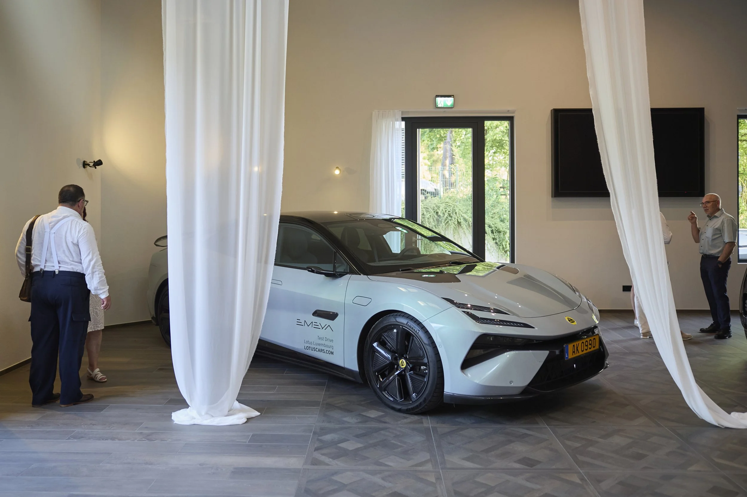 Voiture électrique blanche exposée dans une salle avec quelques personnes. La voiture porte un logo Lotus et la plaque d'immatriculation AK 0990.