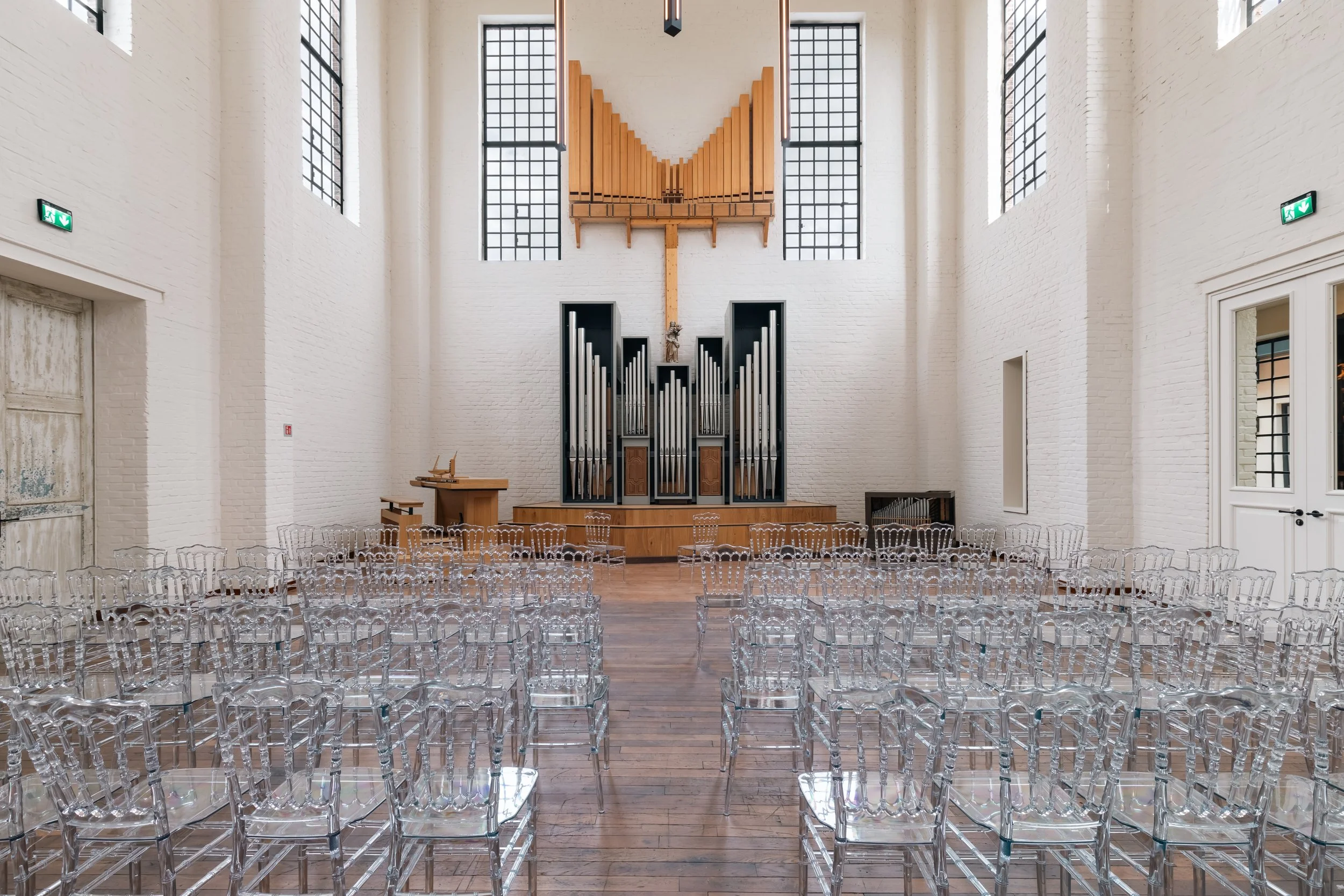Salle de cérémonie avec sièges en plastique transparent, orgue et foyer en bois dans une église moderne avec murs en briques blanches et grandes fenêtres.