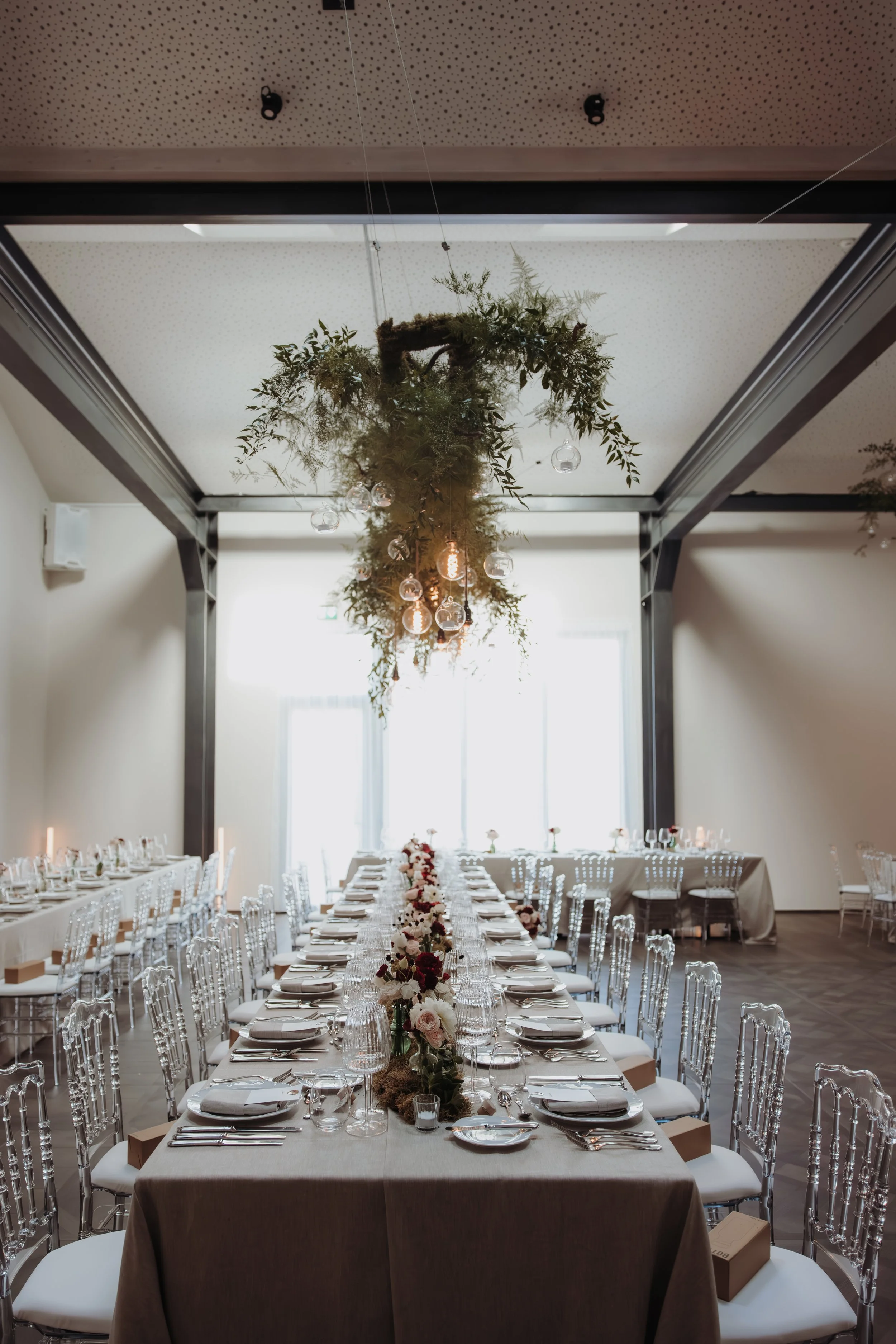Salle de réception élégante avec une longue table décorée de fleurs, verre à vin, assiettes et couverts, et un éclairage suspendu en forme de cascade de plantes et de bougies.