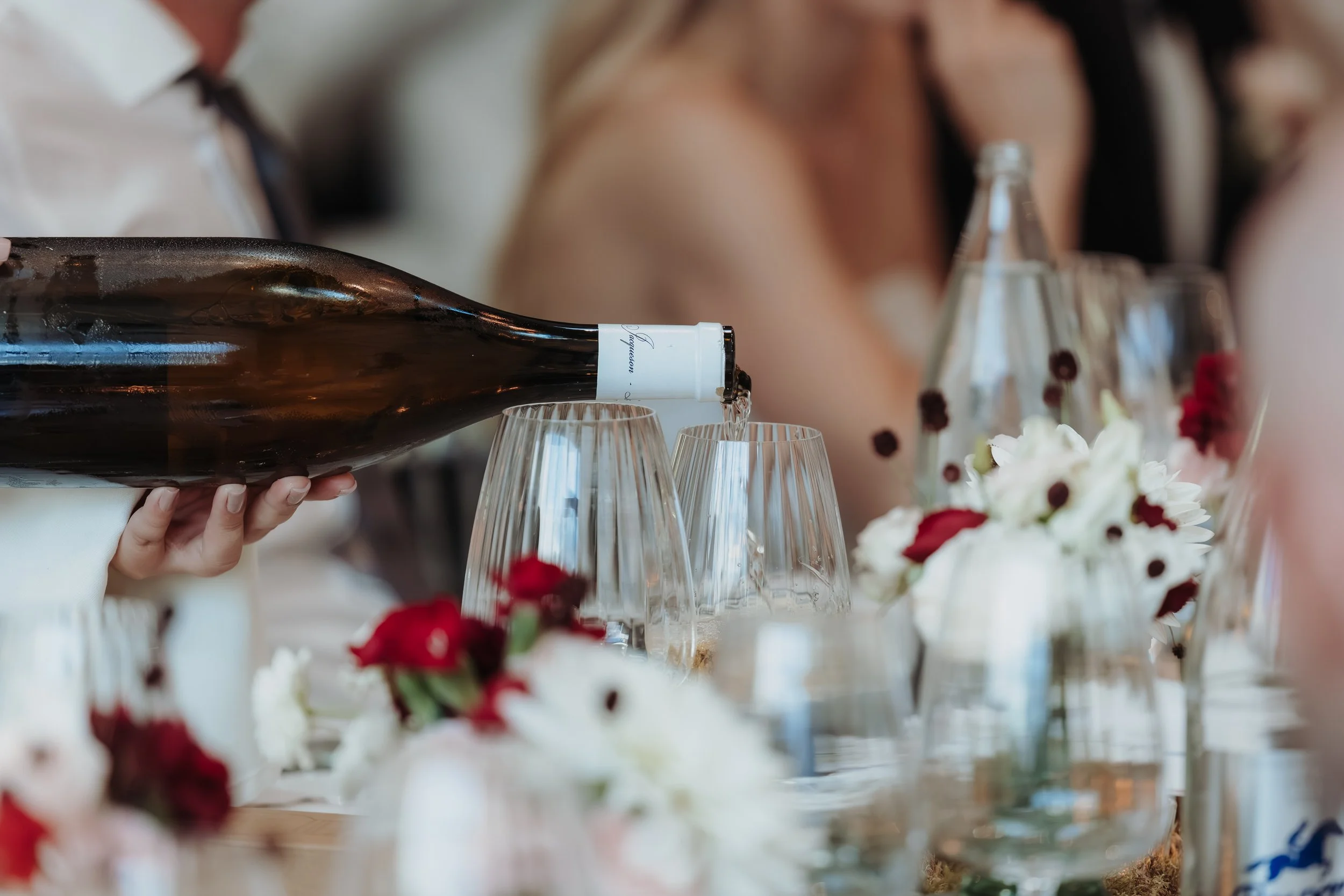 Une personne verse du vin dans des verres lors d'un repas. La table est décorée avec des fleurs blanches et rouges.