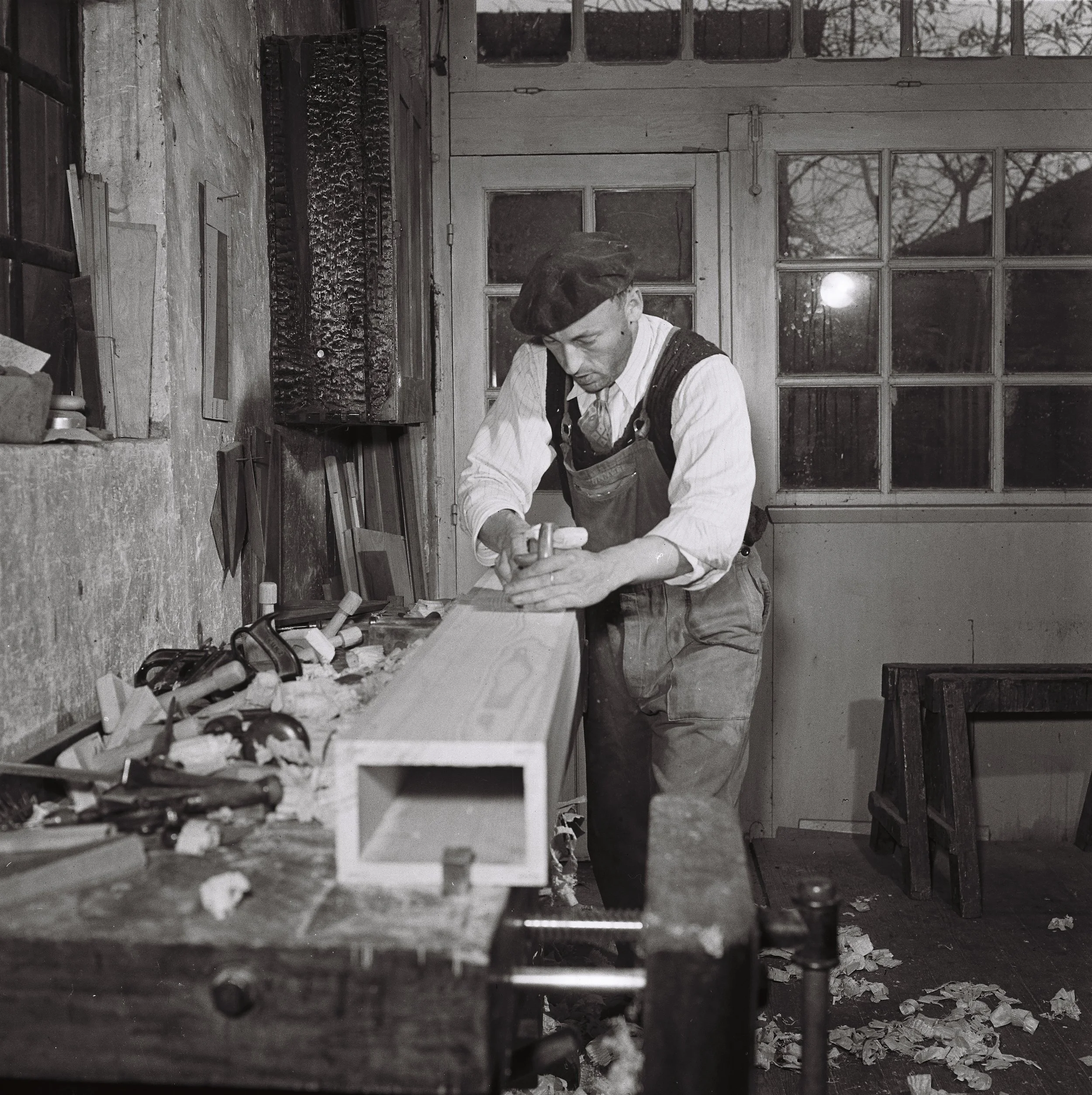 Un homme travaillant dans un atelier, utilisant une râpe pour poncer un morceau de bois, entouré d'outils et de morceaux de bois, avec des fenêtres en arrière-plan.