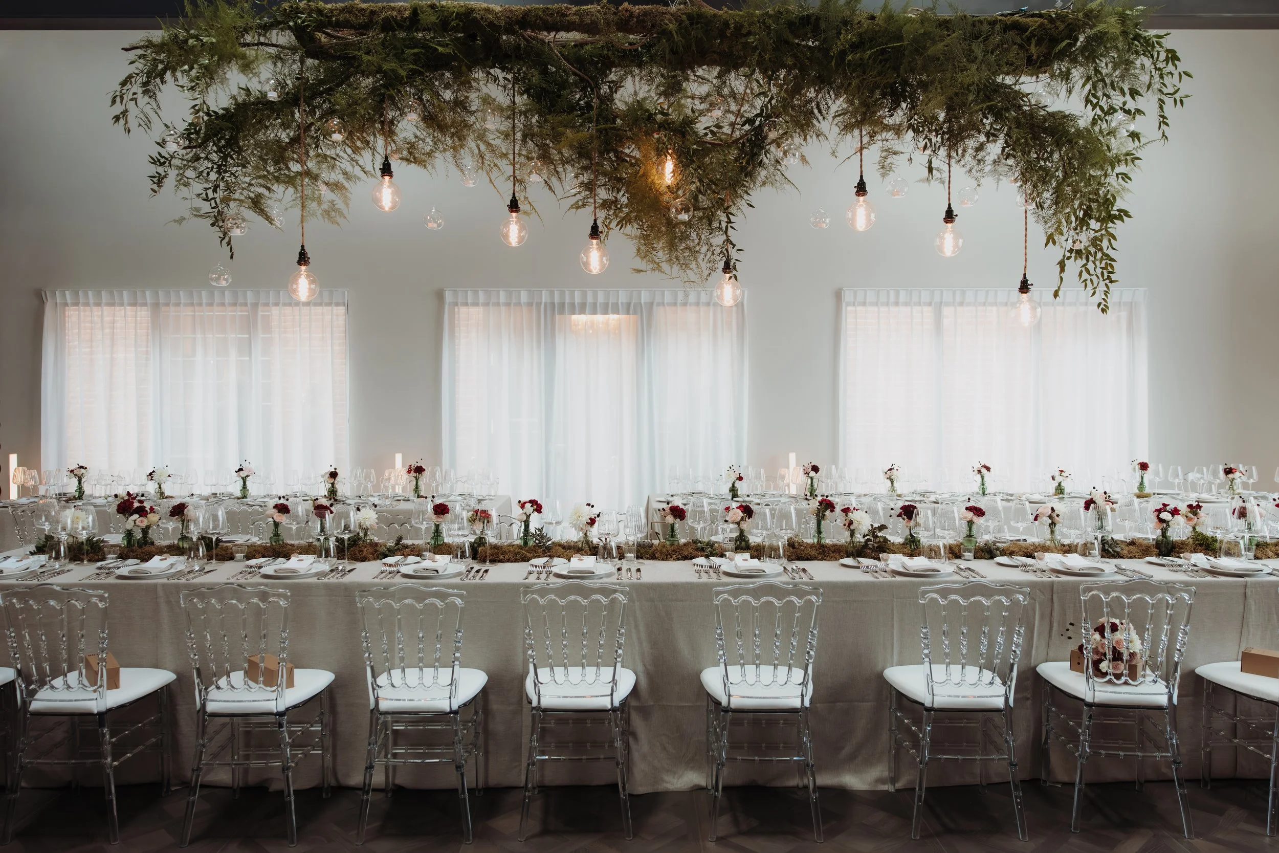 Salle de réception élégante avec un long banquet couvert d'une nappe beige, décorée de petites bouquets de fleurs rouges et blanches, dans une pièce lumineuse avec de grandes fenêtres et des rideaux blancs, un plafond orné de branches vertes et de lampes à ampoules suspendues.