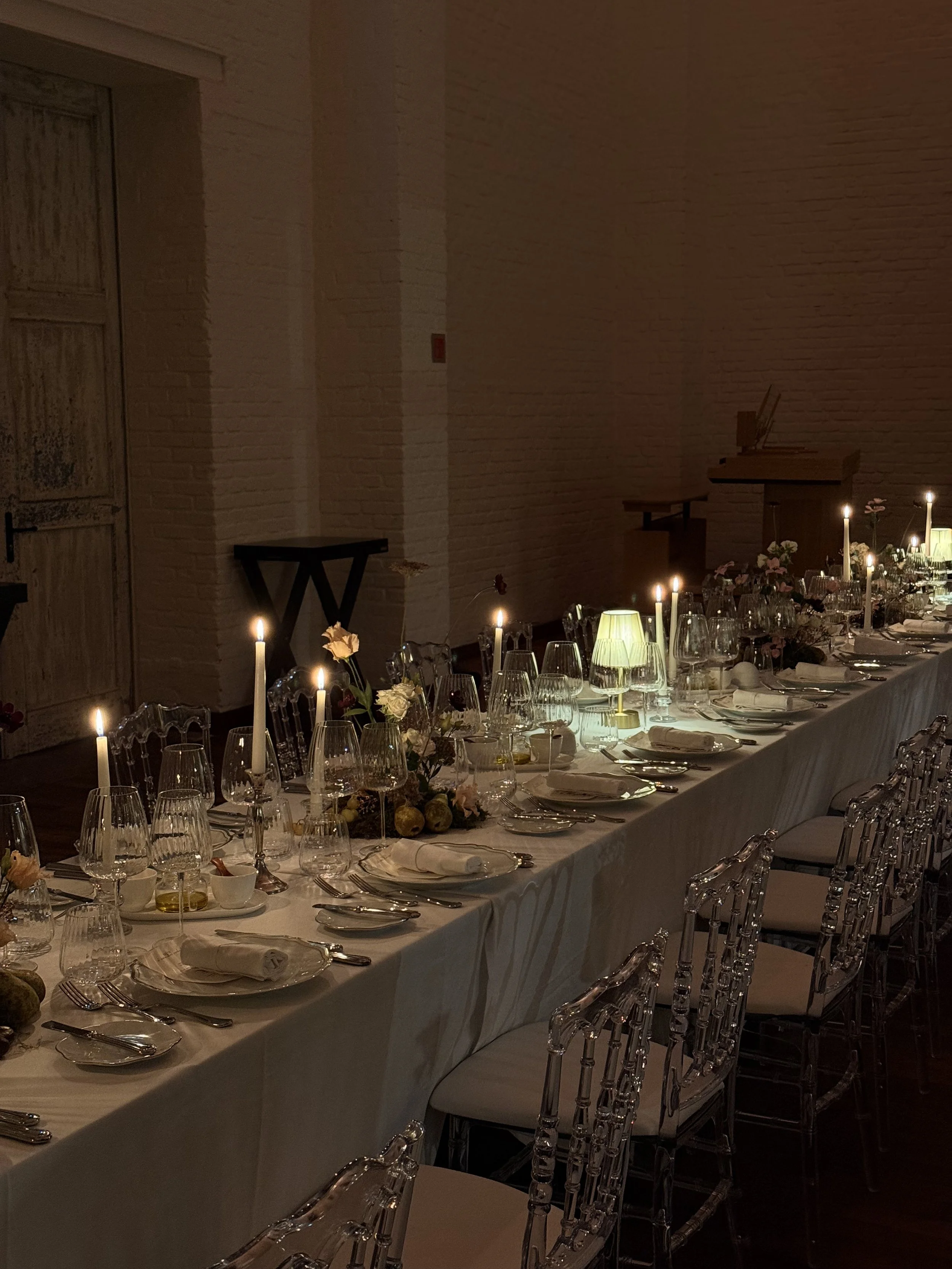 Une longue table de dîner élégamment dressée avec des bougies allumées, des verres à vin, des assiettes en porcelaine, des couverts en argent, et des décorations florales dans un espace intérieur sombre et sophistiqué.