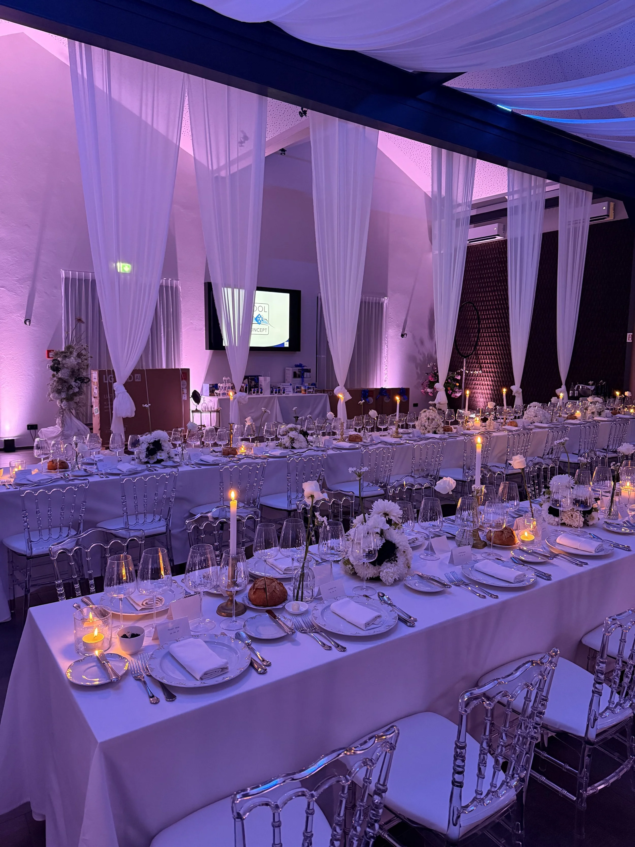 Salle de réception élégante avec longues tables drapées de blanc, décorée de fleurs blanches et bougies, avec un éclairage violet et une toile de fond noire.