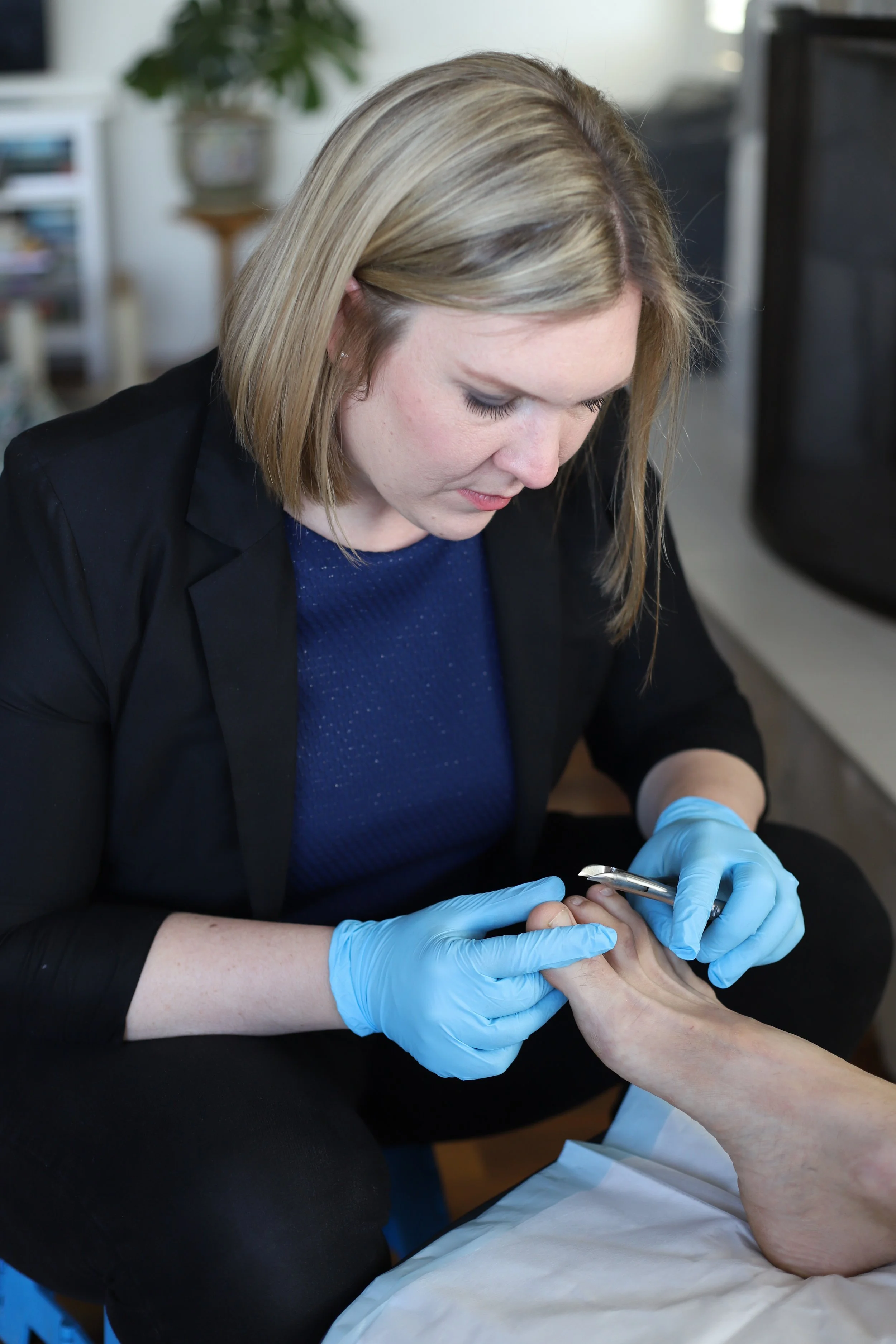 Director and Principal Podiatrist Kristy Outerbridge wearing a black blazer and blue shirt, wearing blue gloves, is trimming toenails of a person's foot with nail clippers, sitting at a table in a well-lit room.